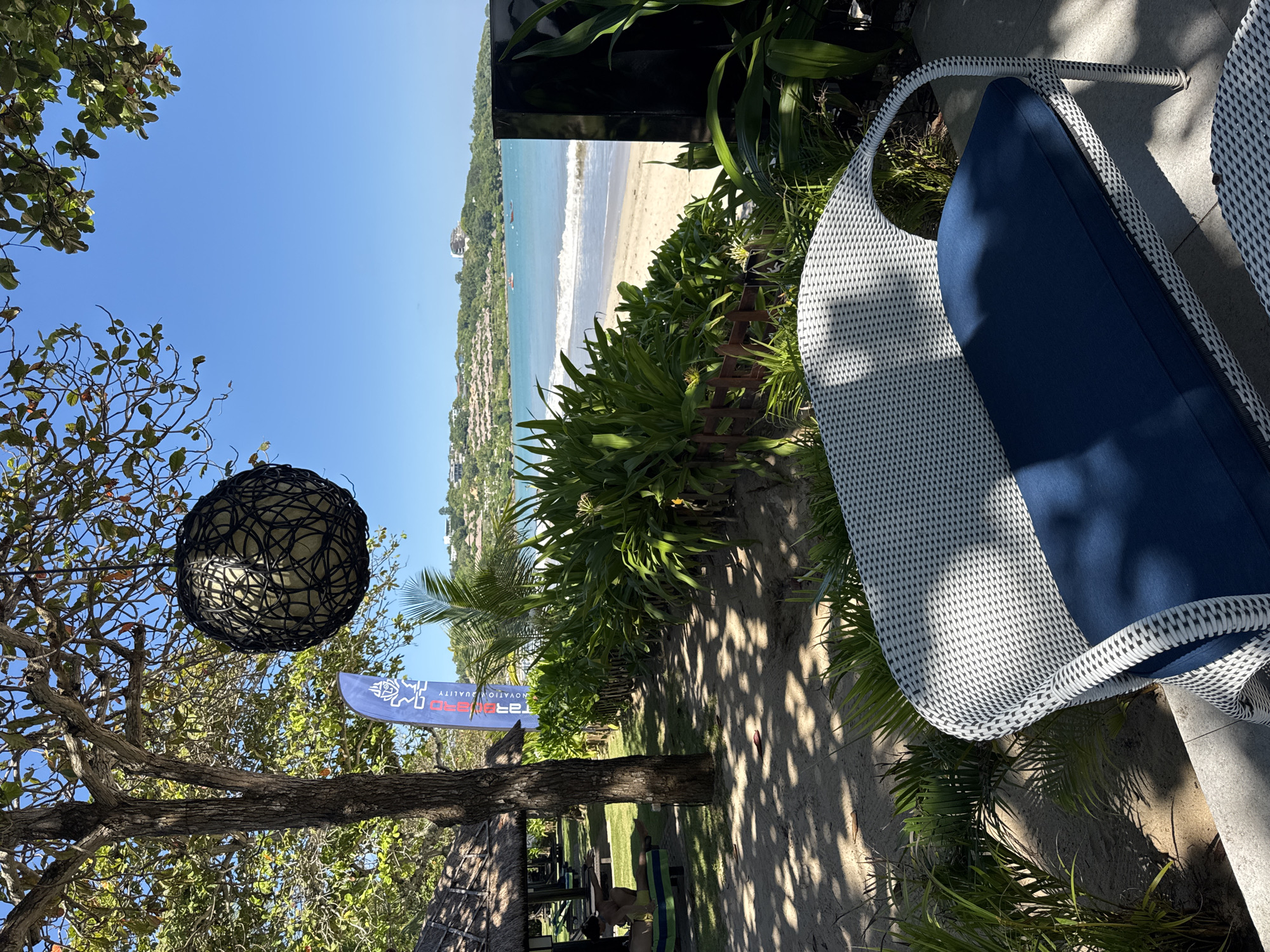 The scene is an outdoor view from what appears to be a patio or elevated area in Kuta Selatan, Indonesia, overlooking a beach. The ocean can be seen in the distance, with a sandy beach stretching along the coastline. A line of buildings and greenery runs along the edge of the beach.
In the foreground, there's a white wicker chair with a blue cushion. The chair takes up a significant portion of the right side of the image. The patio floor is light-colored and appears to be made of stone or tile.
To the left of the chair, dense tropical foliage is visible, including various green plants. A sandy path or area with dappled sunlight is present in the middle ground.
A tree is situated on the left side of the frame, with branches extending toward the sky. Hanging from the tree is a spherical, woven lamp. Below the tree, there are glimpses of people relaxing on sunbeds, under what may be a thatched roof or umbrella structure. A blue banner with white text is also noticeable. The sky above is clear and blue.