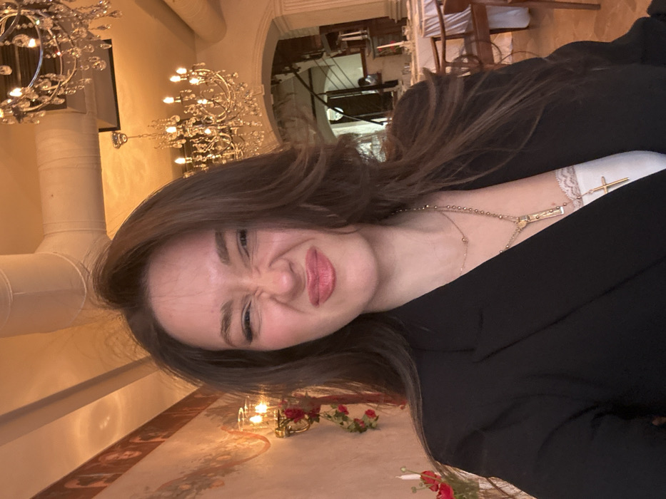 A mid-shot depicts a female subject situated within an indoor dining establishment identified as Florence Restaurant, located in Yerevan, Armenia. The subject, a woman with long, dark brown hair, is centered in the foreground, facing the camera with a distorted facial expression characterized by squinted eyes, a wrinkled nose, and pursed lips. She is dressed in a black blazer worn over a white lace-trimmed top and accessorized with two necklaces, one of which prominently displays a gold-colored cross pendant. The background reveals the restaurant's interior with warm, ambient lighting. To the left, a light-colored wall is adorned with classical murals or frescoes, multiple gold-toned wall sconces illuminating candle-like bulbs, and red floral arrangements. The ceiling features exposed white ventilation ducts. To the right, several ornate crystal chandeliers with numerous lit bulbs hang from the ceiling. An arched doorway leads to a deeper section of the restaurant, where multiple tables are set with white tablecloths and accompanied by wooden chairs.
