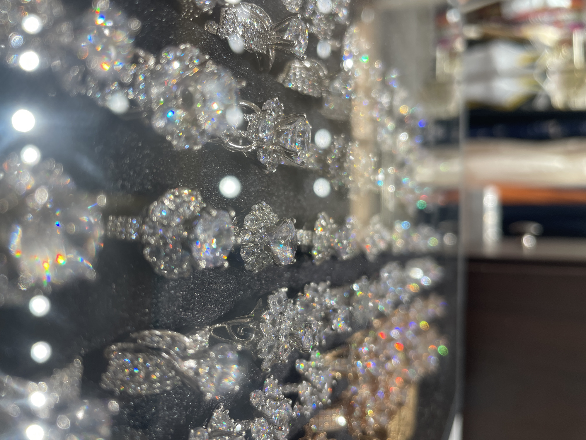 The image shows a display case of sparkly rings, likely made of silver or white gold and adorned with diamonds or cubic zirconia, in Nha Trang Ward, Vietnam. The rings are presented on a dark, possibly black, display pad. The background is blurred but suggests an indoor setting with reflective surfaces. Refraction of light causes rainbow-like sparkles on the objects.
