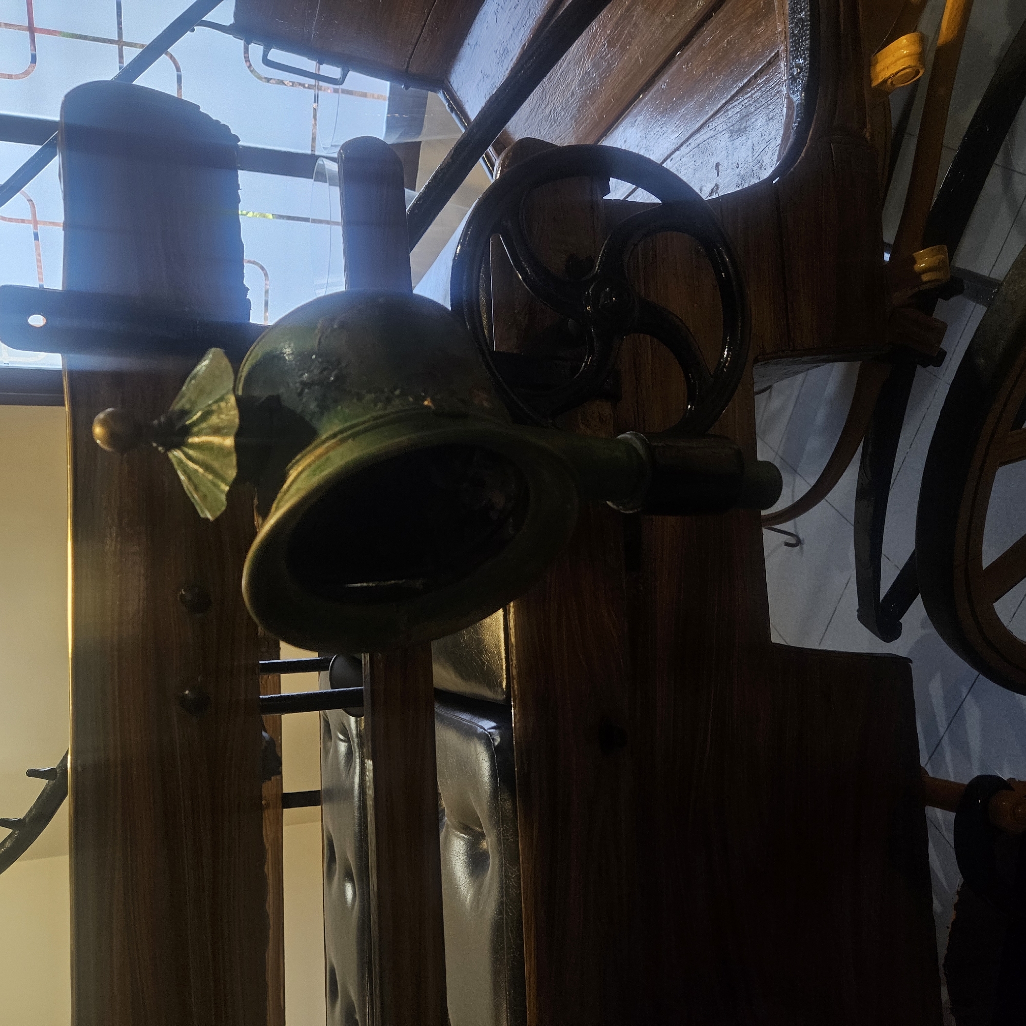 The image shows a close-up view of an antique horse-drawn carriage, likely located in Almadrones, Spain. The carriage is predominantly made of dark wood with leather seats. A prominent object in the foreground is a green, brass-topped horn, possibly a bugle or a signaling device, mounted on the side of the carriage. Attached to the carriage's frame is a black, spoked wheel. The background reveals a patterned window, suggesting an indoor setting, possibly a museum or a private collection. The lighting is dim, creating shadows that add to the vintage feel of the scene.