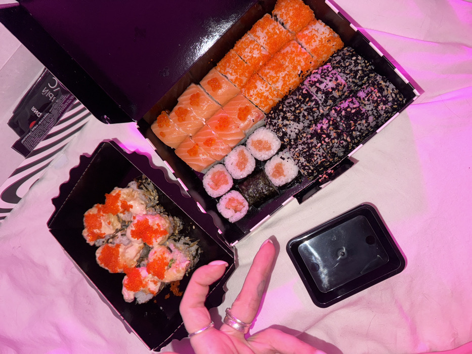The image displays a top-down view of sushi takeout, consistent with delivery in Moscow, Russia. Two open black cardboard boxes containing assorted sushi rolls are positioned on a rumpled white textile surface, illuminated by strong pink/magenta lighting.
The upper-left box contains six large, baked or fried sushi rolls, each topped with a creamy sauce and bright orange tobiko. The lower-right, larger box holds multiple rows of various sushi types:
- A row of eight maki rolls with salmon filling.
- A row of eight rolls topped with sliced salmon and individual orange tobiko.
- Two rows of eight maki rolls entirely covered in bright orange tobiko.
- Two rows of eight maki rolls coated in a mix of black and white sesame seeds.
A left hand, with two silver rings on the ring finger and one on the middle finger, points with its index finger towards a small, empty, black rectangular plastic container, likely for soy sauce. Partially visible in the upper-right corner is a white and black striped paper package, containing a black sachet with Russian text "рий" and "ЧЕРНЫЙ СВС" (likely a sauce packet).