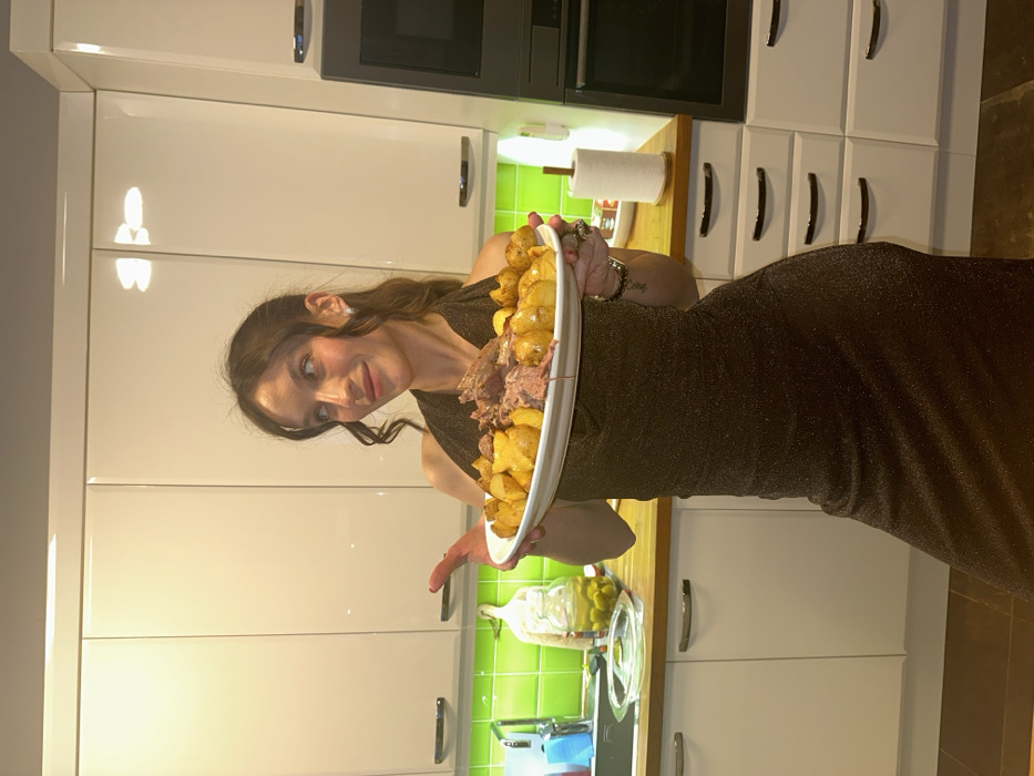 In a kitchen located in Tarasovka, Russia, a young adult female stands facing forward, slightly turned to her left, holding a white oval platter laden with roasted potatoes and a portion of cooked meat. She wears a dark, shimmering, form-fitting halter-neck dress and has long dark hair, visible wavy strands around her face, and a silver earring in her visible right ear. Her head is tilted slightly to her right, and she gazes upward and to her right with a subtle smile.
The kitchen features white, high-gloss upper and lower cabinetry with horizontal silver bar handles. A bright green tiled backsplash is visible above the dark wood-toned countertop, which runs along the left and center of the frame. On the counter to the left, a stainless steel sink with a faucet is present, and a clear glass dish with a lid sits nearby. Further left on the counter, a large glass jar filled with green olives is visible next to a light-colored cutting board. To the right of the woman, a paper towel roll on a wooden holder rests on the countertop. In the background, an integrated oven or microwave unit with a black door is built into the white cabinetry on the far right. Overhead, recessed lighting is reflected in the glossy cabinet surfaces. The floor is dark and indistinct.