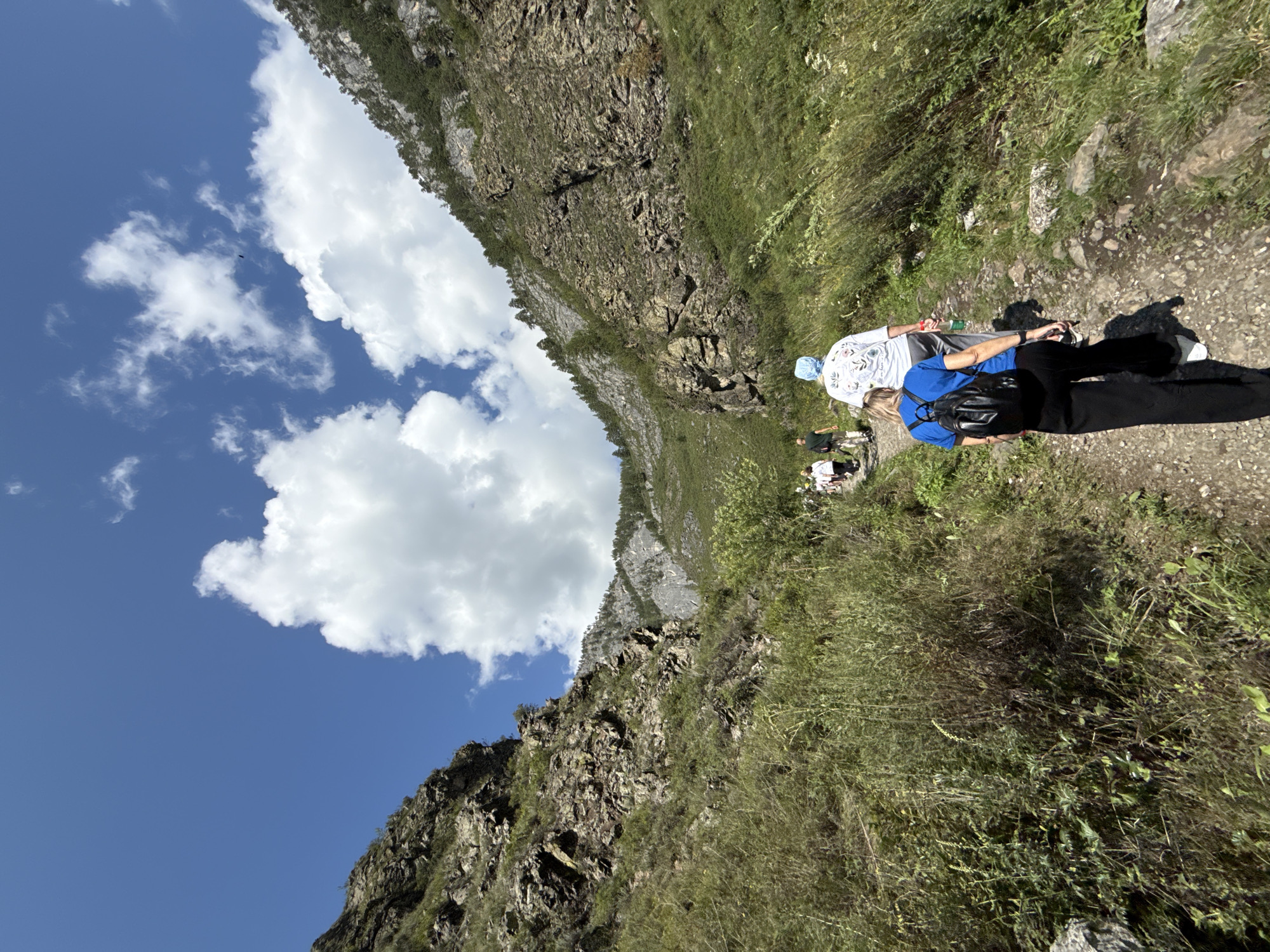 In Chemal, Russia, a group of people are hiking on a rocky, unpaved path through a mountainous, forested area. The path is lined with dense green vegetation and tall grasses. Steep, rocky mountains rise on either side of the path, with sparse trees growing on their slopes. The sky is a clear blue with scattered white clouds. In the foreground, a woman wearing a blue shirt and black pants, carrying a black backpack, walks away from the camera. Behind her, a man in a white shirt and grey pants, wearing a light blue headscarf, walks ahead. Several other hikers are visible further up the path. The scene depicts an outdoor activity in a natural, rugged environment.