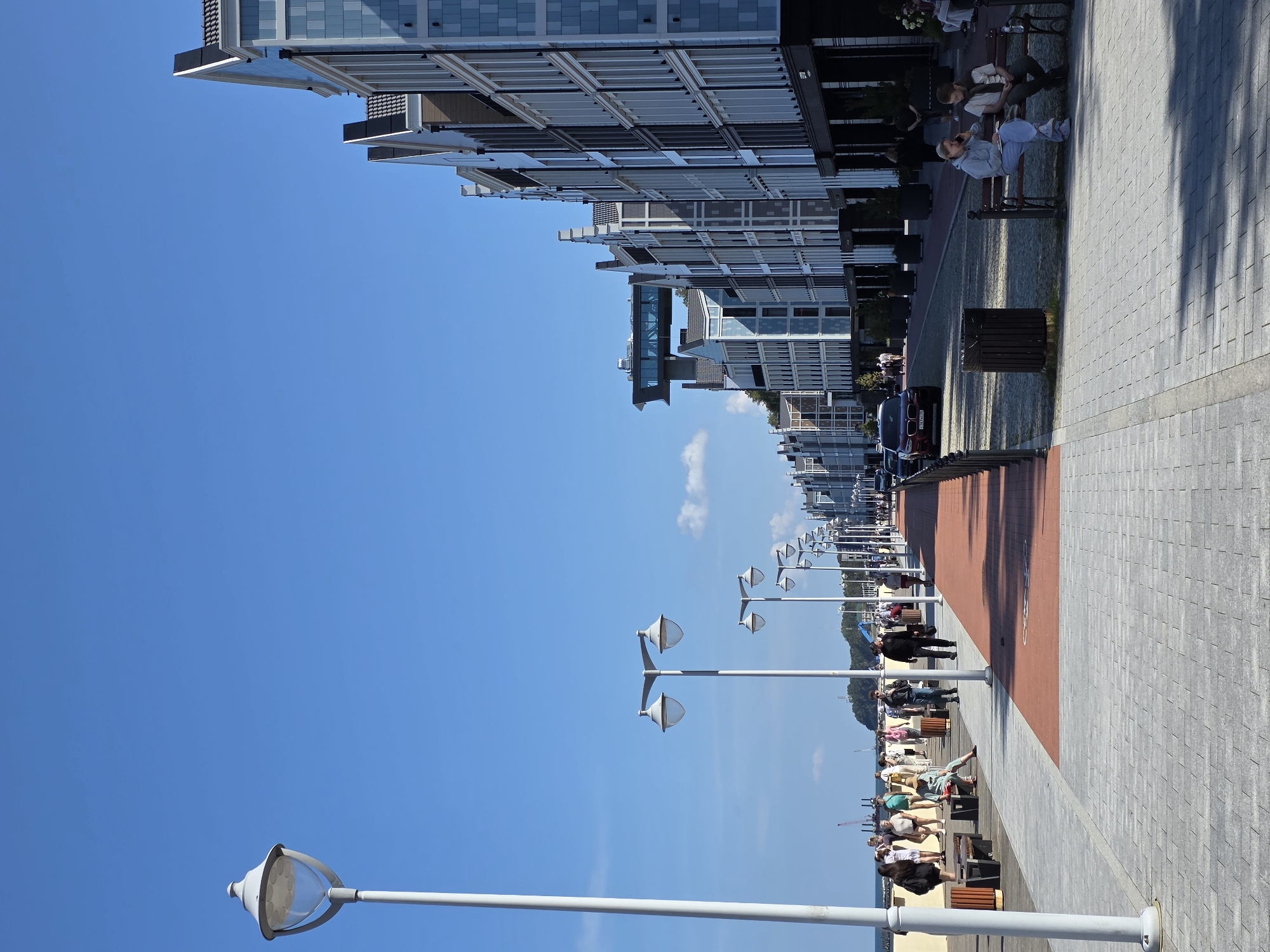 In Svetlogorsk, Russia, a clear blue sky presides over a bustling seaside promenade. The promenade features a paved walkway with a red-colored bicycle lane and is lined with lampposts. On the right side, modern, multi-story buildings with distinct gabled roofs and light blue and white facades stand in a row. Several people are walking along the promenade, some are sitting on benches, and a few cars are parked on the street adjacent to the buildings. The promenade appears to be a popular spot for leisure and recreation, with the buildings suggesting a developed commercial or residential area.