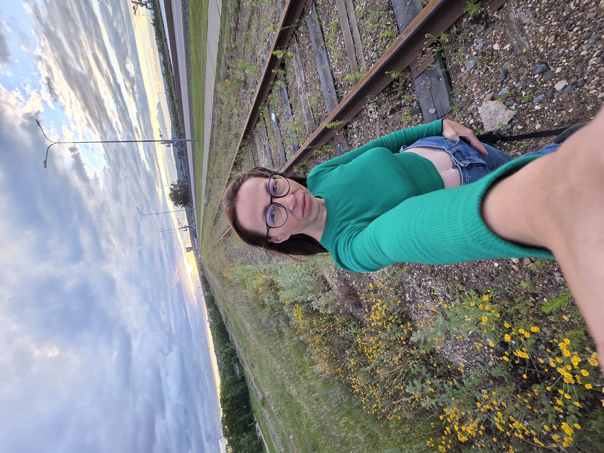 A woman with shoulder-length brown hair and glasses is taking a selfie in Chaikovsky, Russia, near old train tracks. She is wearing a green long-sleeved crop top and blue jeans. The train tracks are overgrown with grass and small yellow flowers. In the background, there is a road, streetlights, trees, and a cloudy sky with the sun setting.