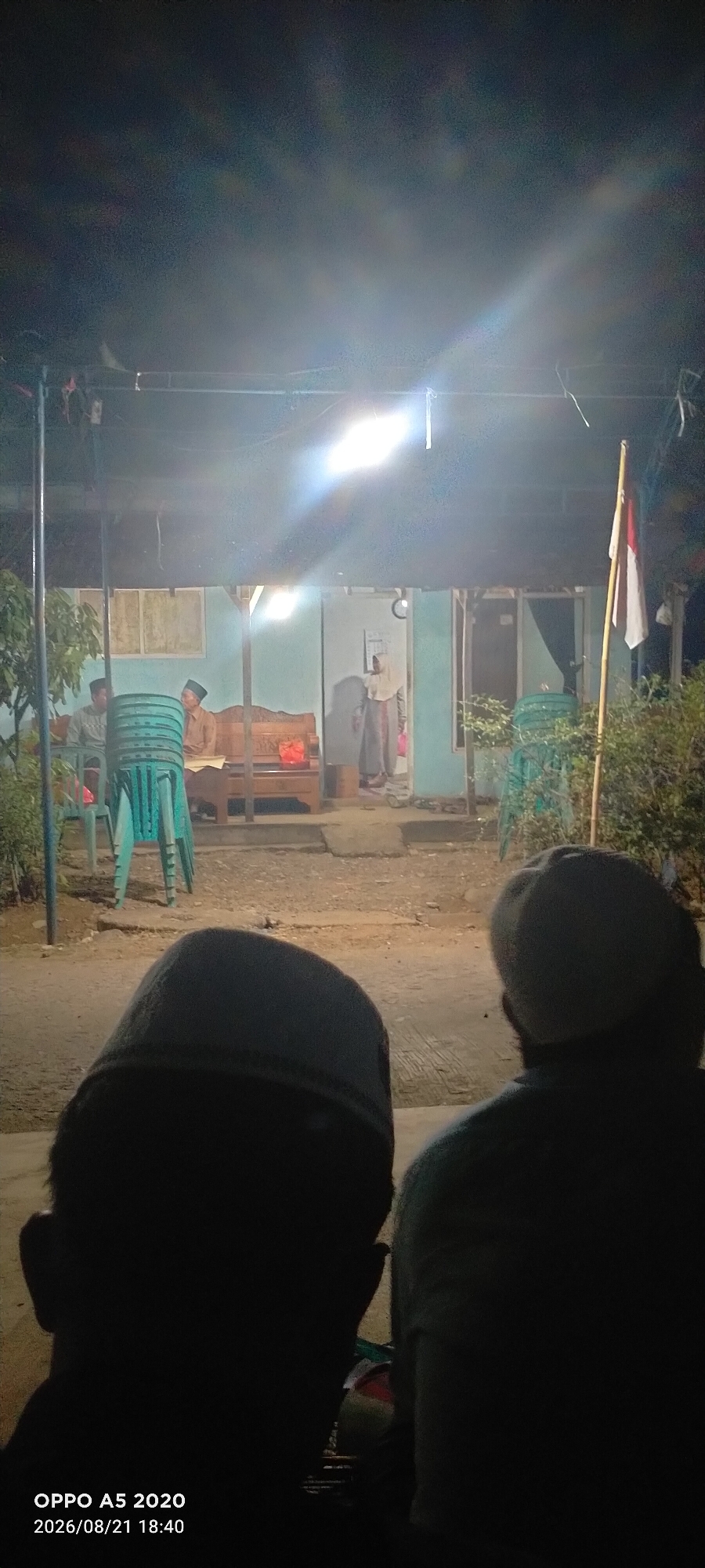 This is a low-light, night-time photograph taken from behind two individuals who are facing away from the camera. The individuals are wearing what appear to be traditional Muslim skullcaps. In the background, a building with a light-colored exterior is visible, illuminated by a bright overhead light. Several people are inside or near the building, some seated on a wooden bench and others standing. There is a large blue water dispenser to the left of the building. A flag, appearing to be the Indonesian flag (red and white), is visible on a pole to the right of the building. The ground in front of the building appears to be dirt or gravel. The photograph includes a timestamp in the bottom left corner indicating "OPPO A5 2020 2026/08/21 18:40". Based on the available information, the location is Sowan Lor, Indonesia. The scene depicts a gathering of people at night, possibly a community event or religious service, in an outdoor setting.
