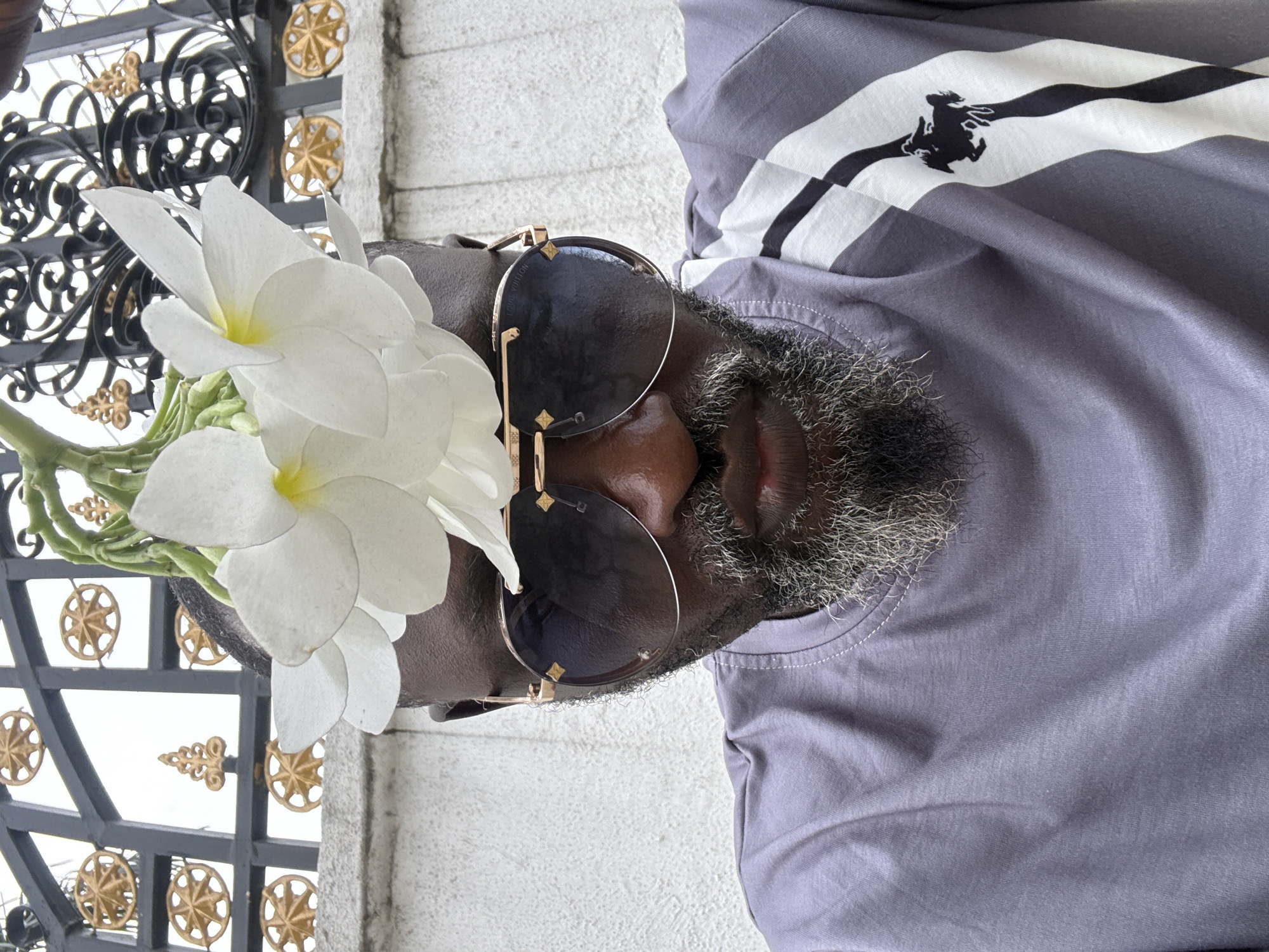 A medium, close-up shot shows a person wearing sunglasses and a grey t-shirt with black and white stripes posing with a bouquet of white flowers. The person has a gray beard. The background consists of a light colored wall and an ornate black metal fence with gold accents. The location is presumed to be within the city of Ado, Nigeria.