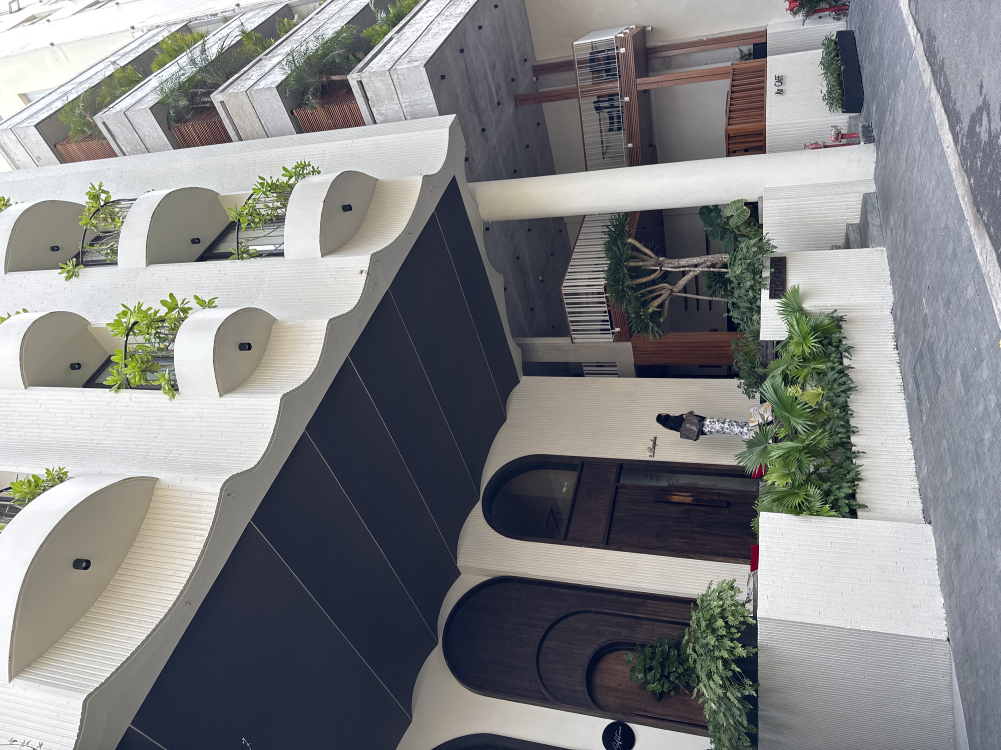 The image depicts the exterior of a modern, multi-story building in Đà Nẵng, Vietnam, observed from a low-angle street-level perspective. The building exhibits a contemporary architectural style characterized by a textured, ribbed white facade contrasting with dark brown and black accents.
The left side of the building features multiple floors with curved, white balconies adorned with black railings and vibrant green potted plants. A large, dark brown overhanging canopy extends from the upper structure, sheltering the ground-level entrance. Below this canopy, two prominent arched wooden double doors serve as the main entry points, with a circular black sign bearing "Cafeteria" affixed to the left of the leftmost door. Lush green foliage is present in planters in front of the entrance. A single individual, a woman, is visible near the entrance, seen from behind/side, wearing a dark top and patterned light-colored trousers, carrying a dark bag over her left shoulder. She appears to be standing or walking near the entrance.
The right side of the building features a different architectural section with concrete balconies, some incorporating wooden slatted facades and railing details. These balconies also contain extensive greenery, including palm-like trees, and several open-air seating areas with chairs and tables are discernible on the lower levels. A large white supporting column is visible on the ground floor, leading to elevated sections. On the ground level, a white wall with the text "Air Cafe" is visible, along with a red fire hydrant and dark planter boxes containing plants. Another sign reading "The Gly" is affixed to a planter near a set of steps. The ground beneath the building consists of dark grey paver tiles, leading to a segment of asphalt road in the foreground.