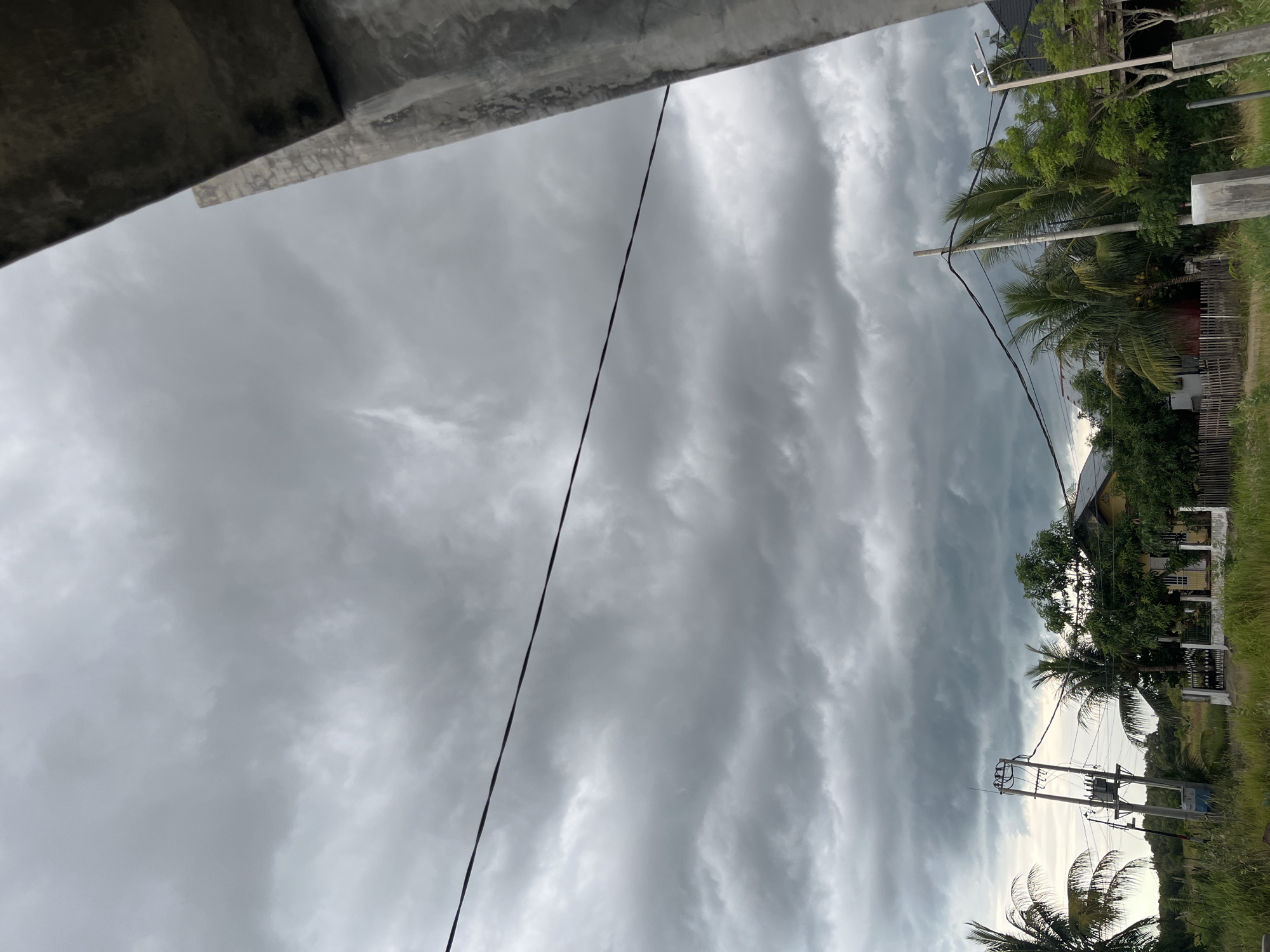 An outdoor, upward-angled shot from Lhokseumawe, Indonesia, captures a predominantly grey, overcast sky. The image is oriented vertically, with a large portion of the frame filled by thick, layered grey clouds that suggest either an approaching storm or recent heavy rainfall.
In the upper left and extending into the upper center, the underside of a concrete architectural element, possibly an overhang or roof edge, is visible. From this concrete structure, a single dark electrical wire or cable descends diagonally across the cloudy expanse towards the lower center of the frame.
The right side of the image reveals a vibrant section of a semi-rural landscape. Dense tropical vegetation, including prominent palm trees and other leafy trees, forms a green backdrop. Interspersed within this greenery are multiple utility poles, some equipped with electrical wires and what appears to be a transformer. A house with a dark roof and lighter walls is partially visible through the foliage and behind a wooden fence towards the upper right. Further to the right, patches of green grass or fields are visible.
No people or discernible activities are present in this static scene. The diffused light from the heavy cloud cover indicates it is daytime. There is no visible text in the image.
