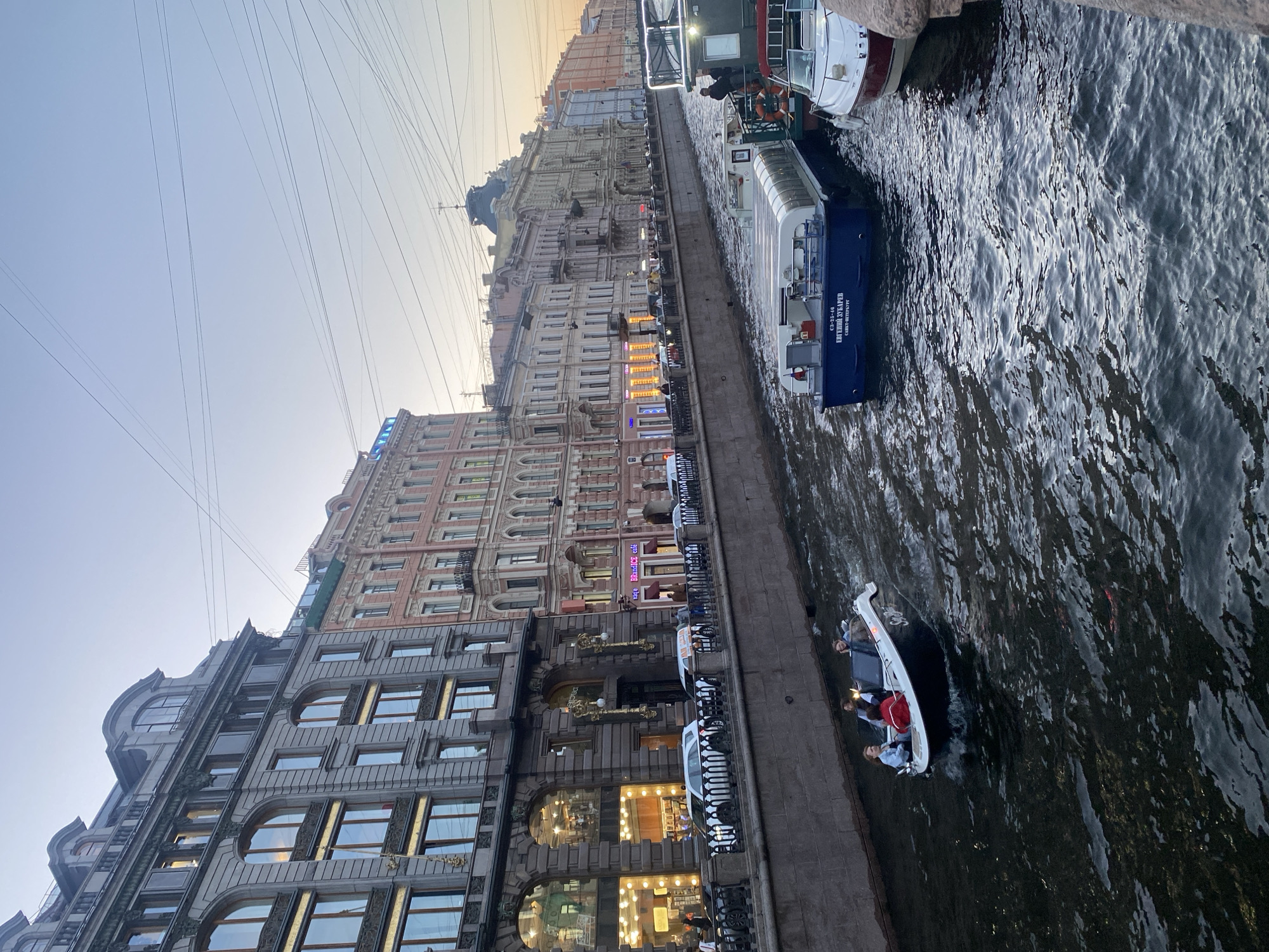 A canal-side view of St. Petersburg, Russia, features a waterway lined with ornate, historic buildings. Several boats are visible on the canal. In the foreground, a small, dark-hulled boat with a white interior is navigating the water, carrying four passengers. Further down the canal, a larger, covered tour boat, primarily blue and white with a glass-enclosed cabin, is docked. To its right, a smaller white boat with a red stripe is also docked. The buildings along the canal are multi-story structures with detailed facades, displaying a variety of architectural styles. The scene suggests a leisurely day in the city, with the buildings illuminated by warm interior lights and the sky displaying a soft twilight glow.