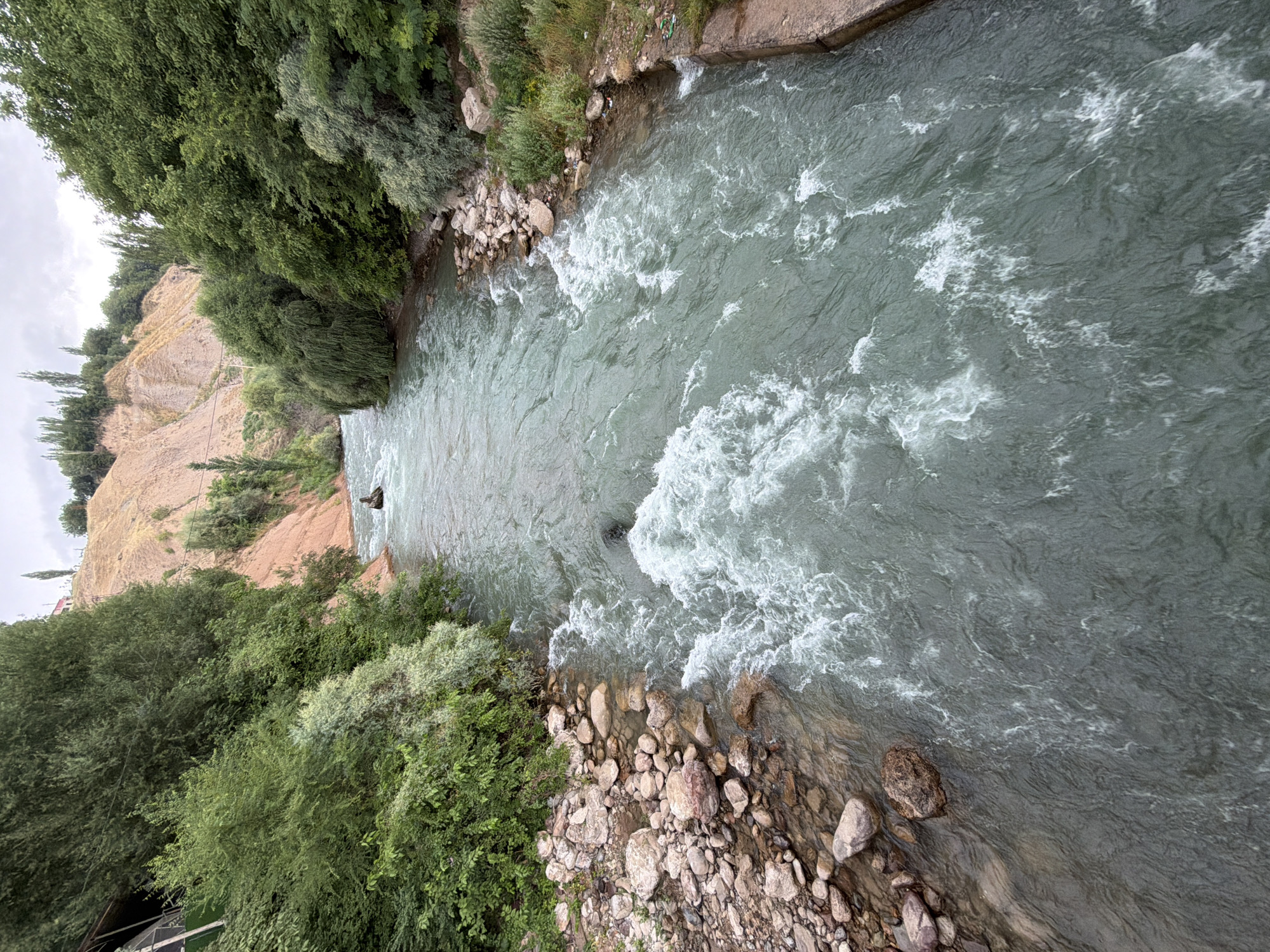 The image shows a wide, fast-flowing river with turbulent water. The river is surrounded by lush green trees on both banks, with a steep, sandy hillside in the background. In the middle of the river, a person is visible in a dark object, appearing to be navigating the rapids. On the left bank, there is a small, covered structure with a green roof. The riverbed appears to be rocky, with stones visible along the edges. The sky is overcast, suggesting a cloudy day. The scene depicts a natural environment with a recreational activity taking place. The location is identified as Khumsan, Uzbekistan.