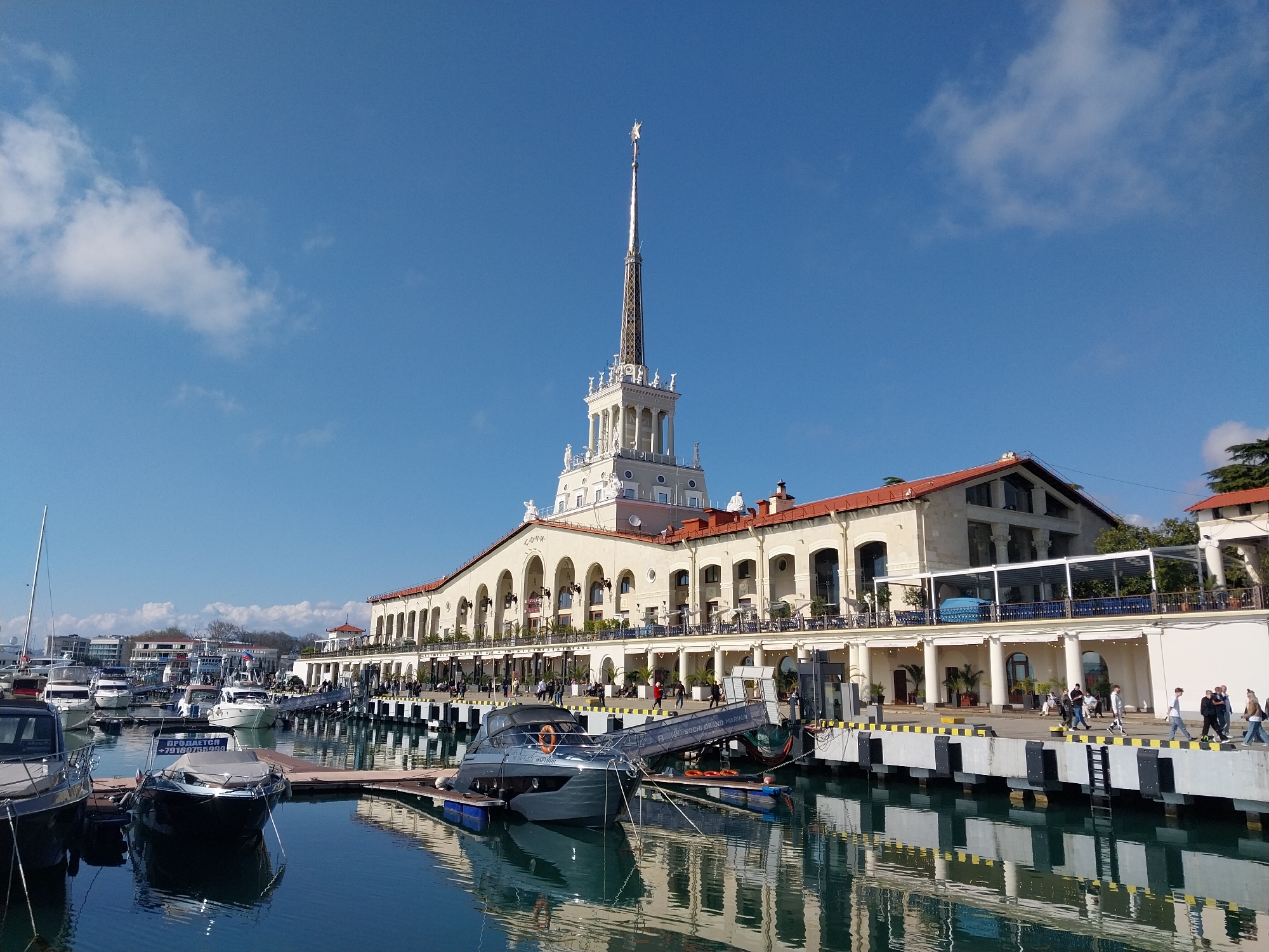 This daytime image showcases the grand Sochi Seaport building and its bustling marina in Sochi, Russia, under a clear blue sky with scattered white clouds. The prominent building, an example of Stalinist Empire style, features a light-colored facade with numerous arched windows and balconies, a red-tiled roof, and a tall, elegant central spire topped with a shining star or ornate element. Statues can be seen adorning the spire's base. The word "СОЧИ" is faintly visible on the building's facade.
In the foreground and mid-ground, a marina is filled with numerous boats and yachts of varying sizes, docked along black and yellow striped piers. The calm water beautifully reflects the building and boats, creating a serene effect. People are seen strolling along the waterfront promenade, some gathered near the boats, while others appear to be seated at outdoor cafes and restaurants located on the building's terraces.
Visible text on a boat reads "ПРОДАЕТСЯ +79180775988" (FOR SALE with a phone number), indicating a boat is for sale. Another piece of text on a gangway leading to a yacht reads "ЯХТ КЛУБ SOCHI GRAND MARINA" (YACHT CLUB SOCHI GRAND MARINA), confirming the location as a major marina. The bright sunlight suggests pleasant, warm weather, contributing to the lively and relaxed atmosphere. The scene portrays a popular recreational and tourist destination.