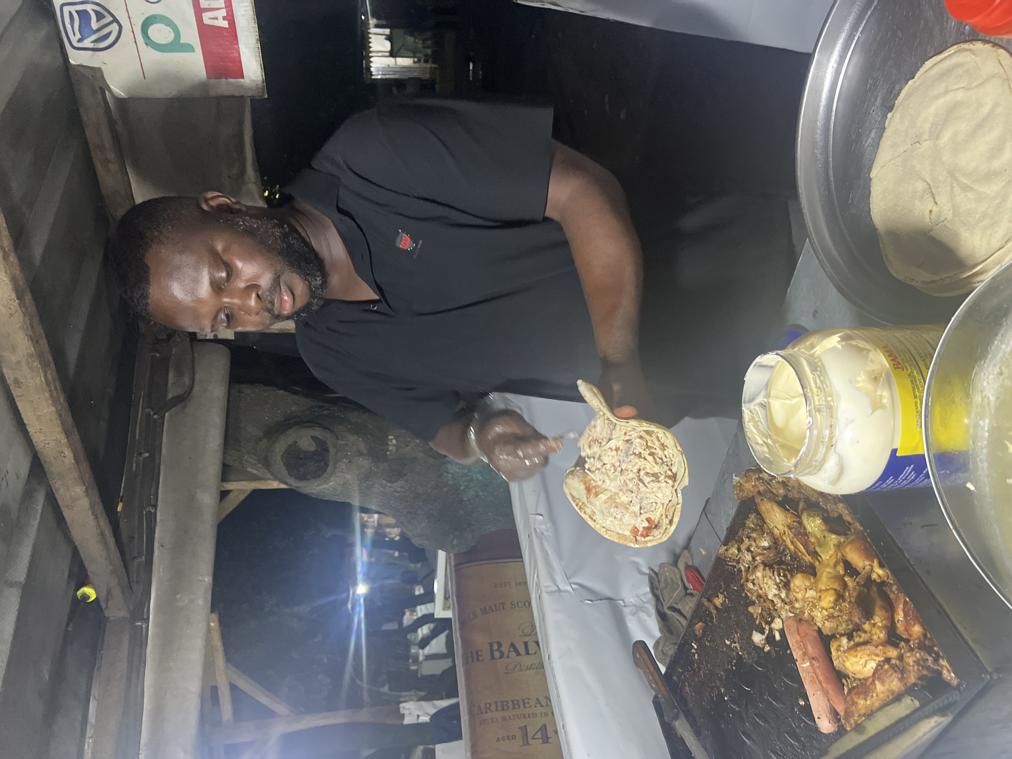 The image captures a close-up scene of a man preparing street food at night, likely in Abuja, Nigeria. The central subject is a dark-skinned man with a beard, wearing a black short-sleeved shirt with a small red and green logo on the chest. He is focused on filling a flatbread, possibly a wrap or local bread, with a mixture that appears to be finely chopped meat or tuna salad, along with some sauce. He holds the flatbread with one gloved hand and uses a utensil in the other.
To his right, an open jar of "BAMA mayonnaise" is visible, along with a metal tray holding a flatbread. Below the prepared flatbread, a dark griddle or tray is filled with various cooked food items, including what looks like grilled chicken pieces and a hotdog, suggesting a mixed grill or a variety of fillings are available. A pair of work gloves and a knife are also on this surface.
The setting appears to be an outdoor food stall or makeshift kitchen, illuminated by artificial light, indicating it's nighttime. In the background, to the man's left, parts of a large cardboard box or bottle are visible, with text reading "THE BALVENIE," "SINGLE MALT SCOTCH," "CARIBBEAN," and "AGED 14." Above this, a sign displays a Volkswagen logo and another green logo resembling a 'p' or an 'ATM' symbol, suggesting a nearby business or banking service. The overall atmosphere points to a bustling urban night food scene. The man exhibits focus and diligence in his task.