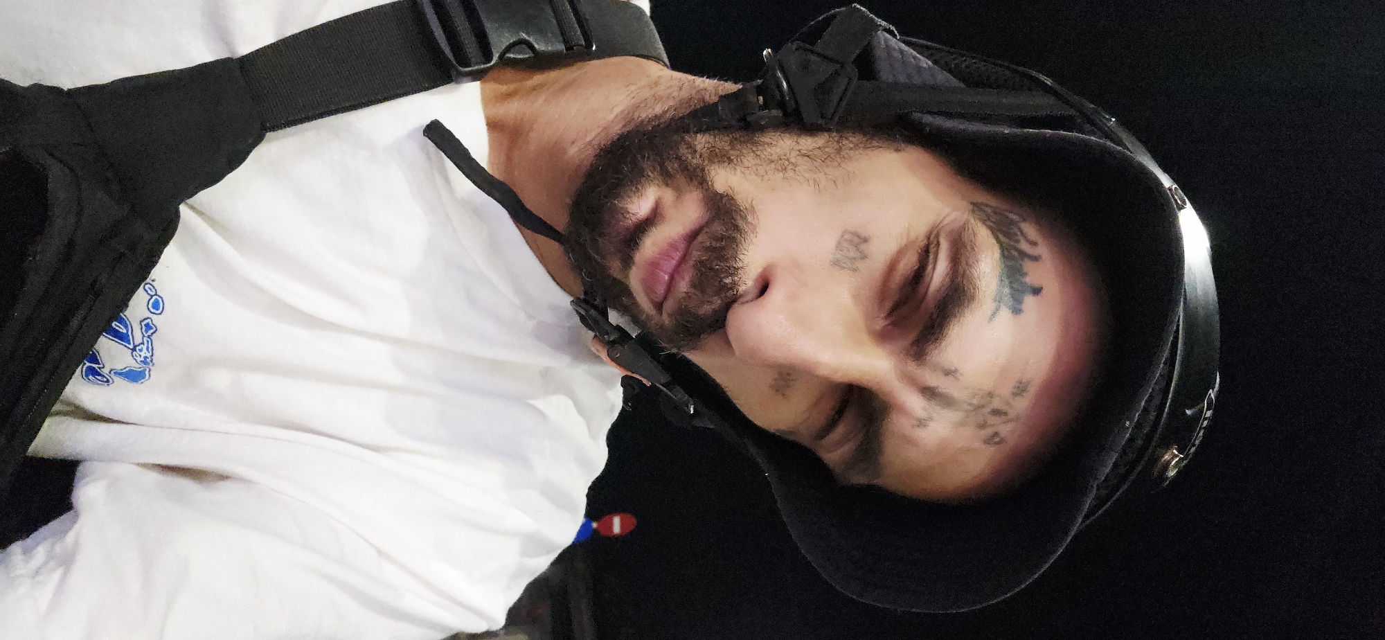 This close-up portrait, oriented sideways, features a man with a beard and prominent facial tattoos, including stylized script on his forehead and a leafy design on his temple. His eyes are closed, and his mouth is slightly open, suggesting he is asleep, resting, or in a state of repose. He is wearing a white t-shirt with a blue and white graphic logo visible on the left side of his chest, and a black harness or strap over his shoulder. A black helmet or protective headgear covers the back of his head and part of his face. The background is uniformly dark, indicating the image was likely captured at night or in a very dim setting, with the subject illuminated by artificial light. The scene conveys a sense of peaceful quiet. The location context is Cam Đức, Vietnam.