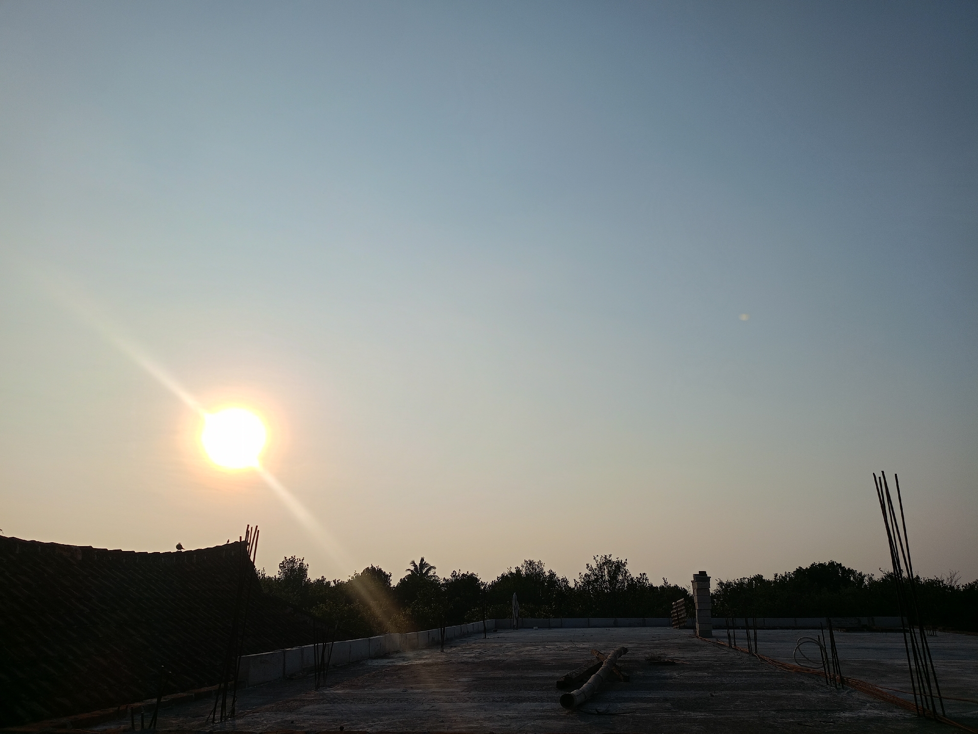 The image depicts a rooftop construction site bathed in the warm glow of a setting sun. The sky is a gradient of pale blue to a hazy yellow, with the sun a bright orb on the left, casting a powerful glare and light rays across the scene. Two small birds are perched on a dark, tiled roof structure in the foreground. The rooftop itself is unfinished concrete, with exposed rebar and construction materials scattered about, including pipes and coiled wiring. A line of concrete walls demarcates the edge of the rooftop, beyond which a dense line of trees forms a dark silhouette against the horizon. The overall impression is one of urban development in a natural setting, likely in Batu, Indonesia, given the subtropical vegetation. The scene is devoid of human activity.