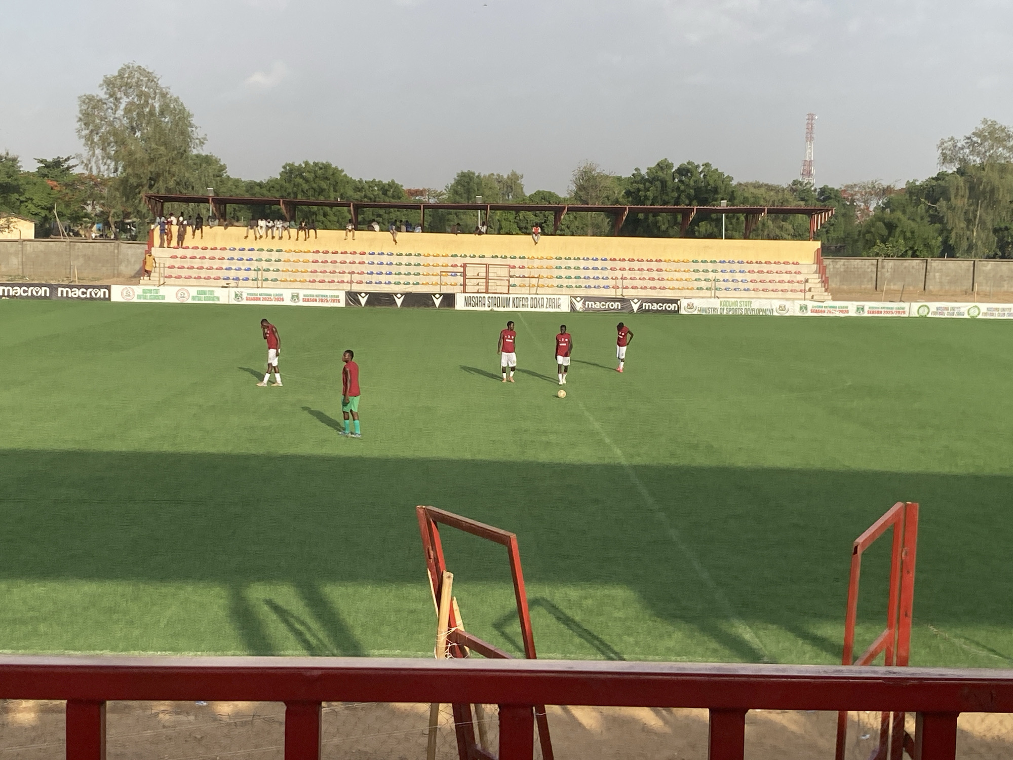 The image shows a soccer field at Nasara Stadium Kofar Doka Zaria, located in Tudun Wada, Nigeria. Five people wearing red shirts with white shorts and socks are visible on the field. One of them is wearing green socks. The field is green and well-maintained. A goalpost, painted red, is visible in the foreground. Behind the field is a stadium stand with yellow seating, where people are sitting. A banner behind the stand displays text and logos, including "NASARA STADIUM KOFAR DOKA ZARIA", "KADUNA STATE MINISTRY OF SPORTS DEVELOPMENT", "SEASON 2025/2026", and the logo of "Macron". A telecommunications tower is visible in the background, along with trees. The sky is overcast.