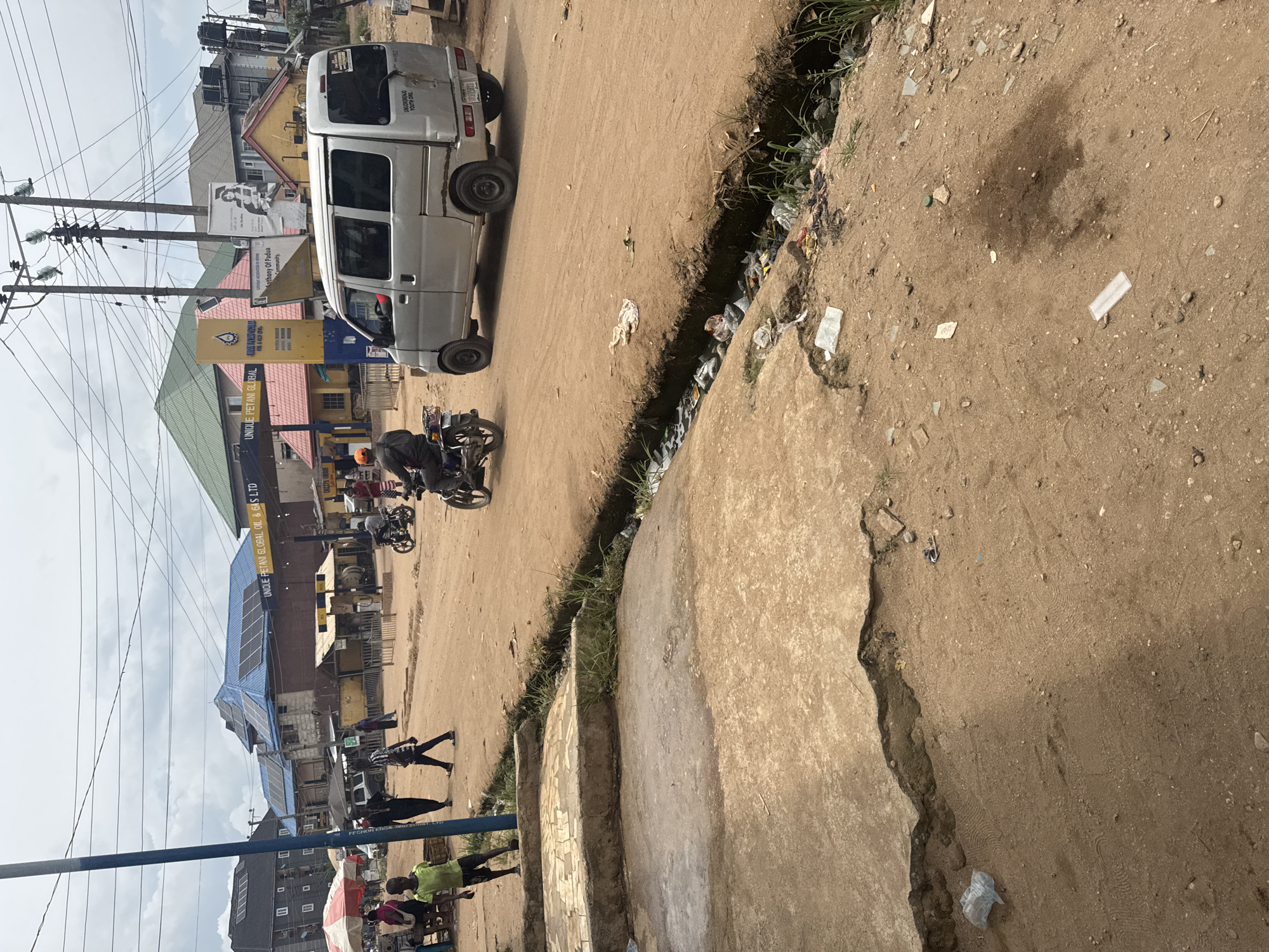 The image depicts a street scene in Ihiagwa, Nigeria, with a van parked on a dirt road next to a concrete curb and a deep, litter-filled ditch. A motorcycle is also present on the road. Several people are walking on the side of the road. In the background, buildings and utility poles can be seen, including a gas station labeled "UNIQUE PETANI GLOBAL OIL & GAS LTD." A sign indicates “Anthony Of Padua Community, and another states “UMUORORONJO YOUTH ORG”.