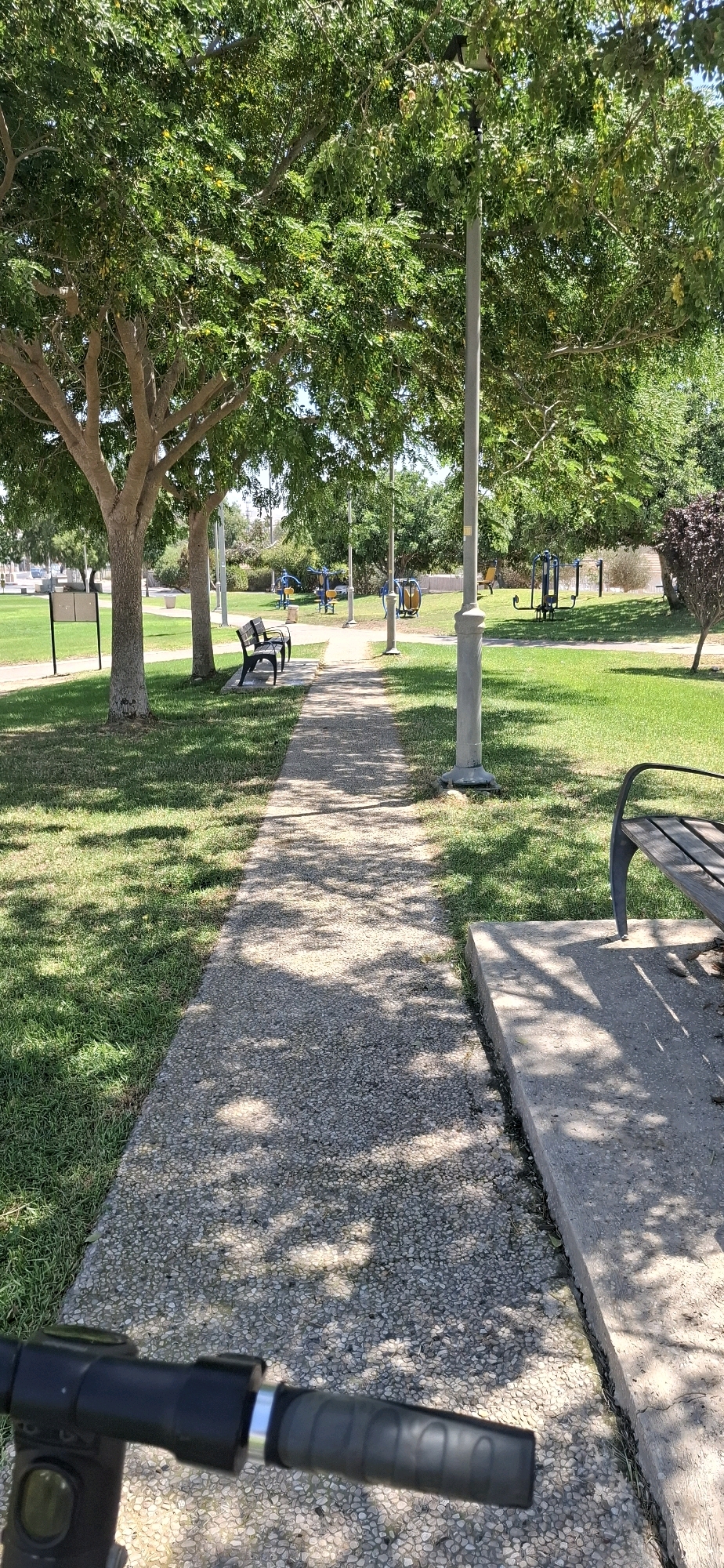 The image depicts a sun-dappled park pathway in Yerocham, Israel. A concrete path, composed of small pebbles, leads into the distance, bordered by manicured green grass and shaded by mature trees with lush green foliage. Several outdoor exercise machines are visible in the background, suggesting a public recreational area. Two dark-colored park benches are strategically placed along the path. In the foreground, the handlebars and part of the frame of a black electric scooter are visible, indicating the perspective of someone positioned on the scooter. The scene is characterized by bright daylight, with shadows cast by the trees creating a mosaic pattern on the ground.