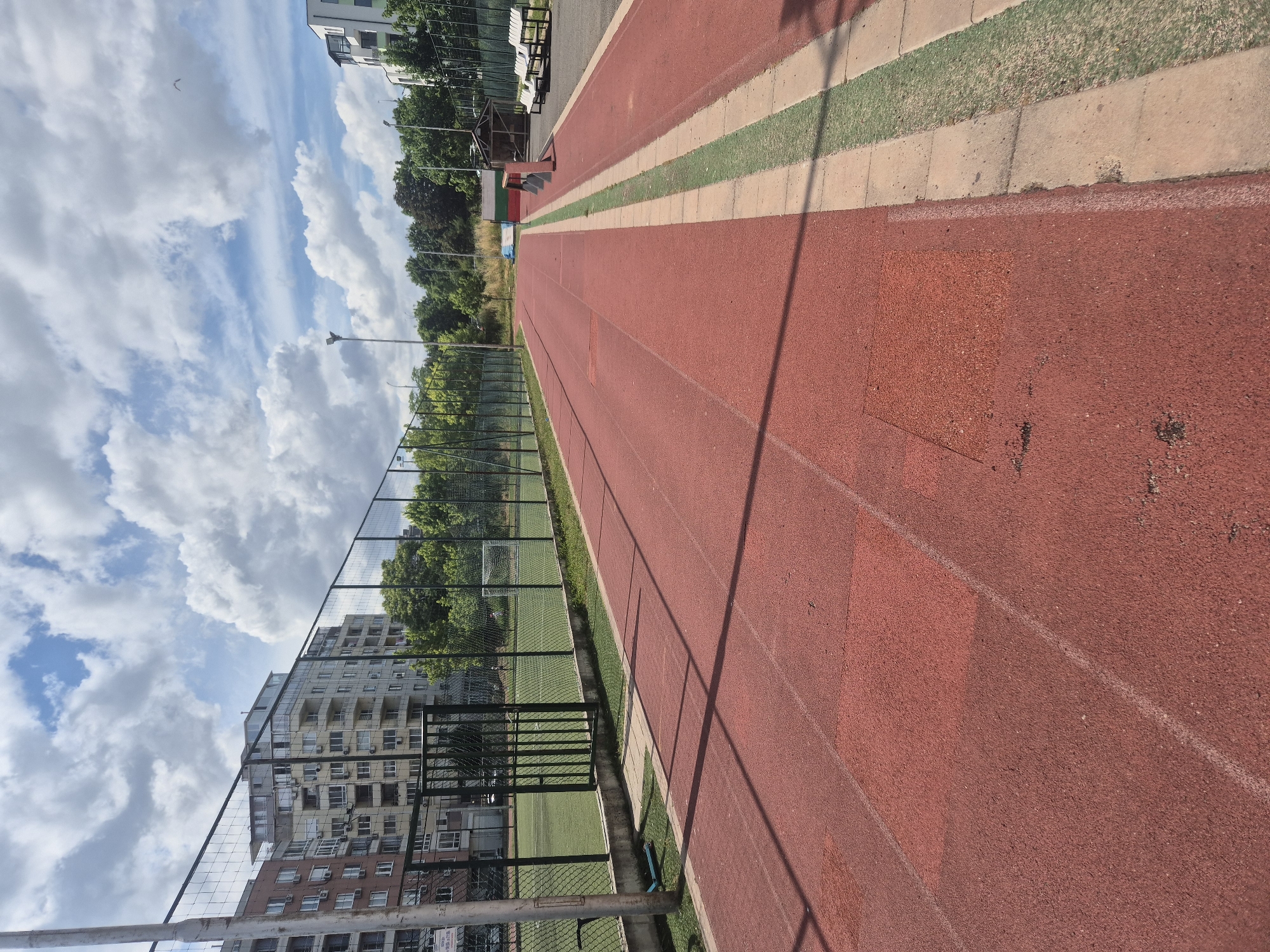 An exterior, eye-level, medium shot captures a red athletic track. The track is smooth, with faint white lines marking lanes and rectangular darker patches of wear. The track runs diagonally from the bottom left foreground toward the middle right background. Beyond the track, a green fence separates it from a soccer field, which is visible on the left. Behind the fence and soccer field are apartment buildings and trees. The sky is visible in the upper half of the image, filled with white, puffy clouds against a bright blue sky. To the right of the track, a green and beige border runs along its edge, with a concrete walkway beyond that. Benches and a small wooden structure are visible further to the right. A bird is flying in the sky in the upper right corner. The scene appears to be a public outdoor sports facility.
