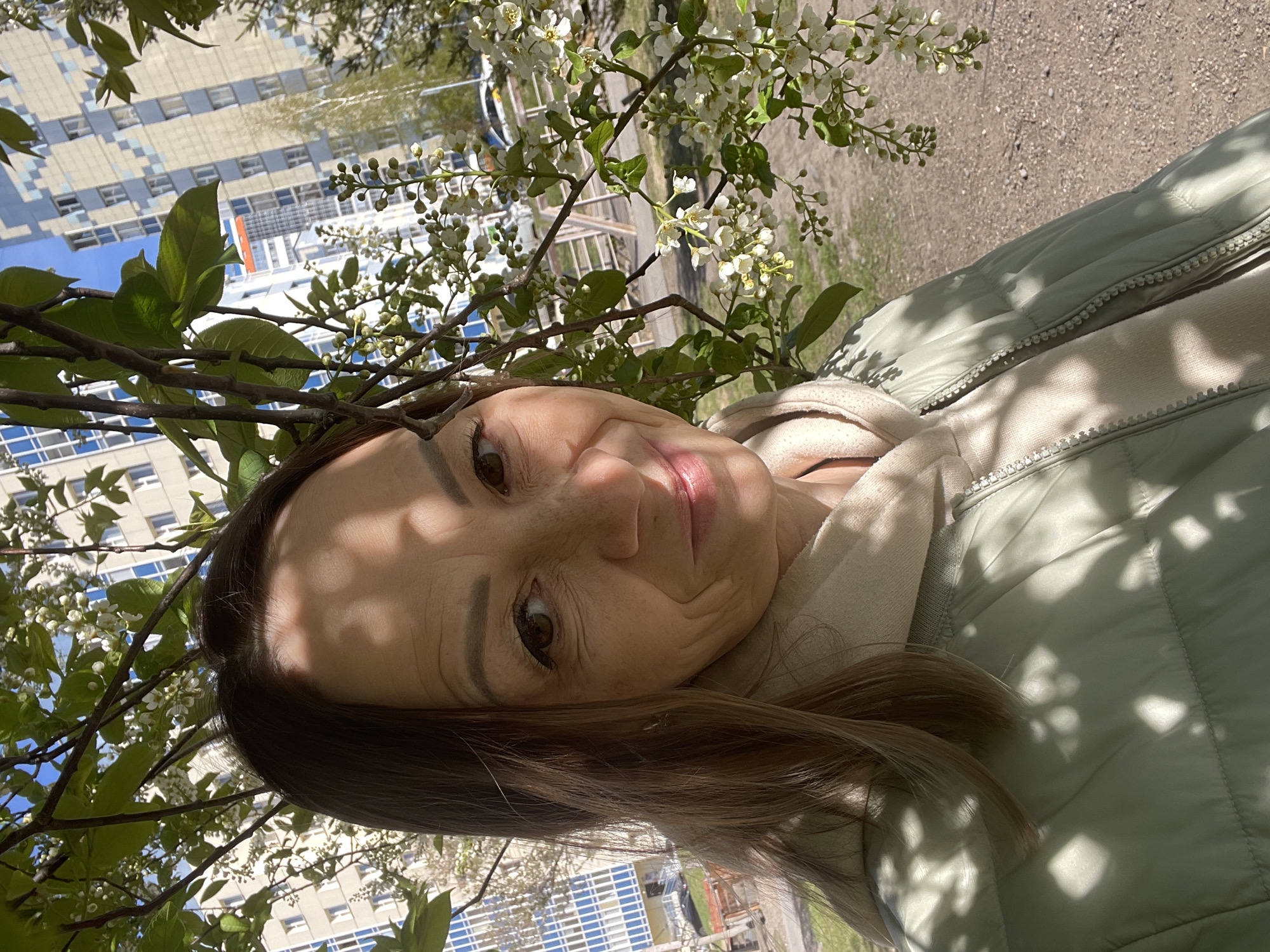 A woman is the main subject of the selfie-style image. She is situated beneath a tree with white blossoms, creating dappled light across her face and clothing. She has brown hair and light skin. She is wearing a light green quilted jacket over a light-colored hooded sweatshirt. The background includes a multi-story building. This photograph was taken in Krasnoyarsk, Russia. The surroundings appear to be an urban environment, with evidence of a paved ground, likely a sidewalk or pathway. The presence of buildings and the style of blossoms suggests it may be spring.
