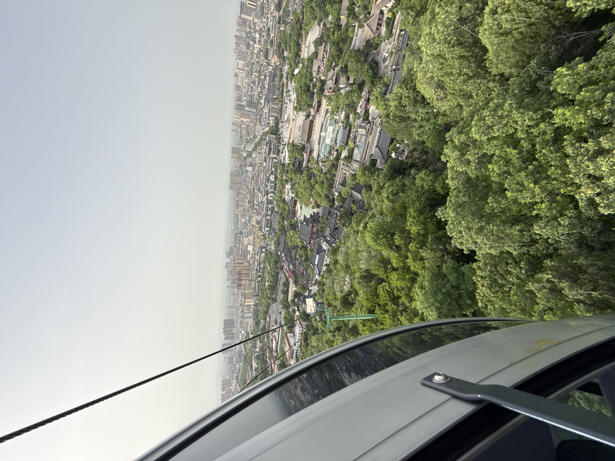 A high-angle shot looking down from what appears to be a cable car reveals a view of Lintong District, China. Dense greenery with trees obscures parts of the city. Buildings of varying heights, from low traditional structures to tall modern apartment blocks, spread across the landscape into the hazy distance. A cable and the roof of the cable car are visible in the foreground. There are no people visible in the scene.