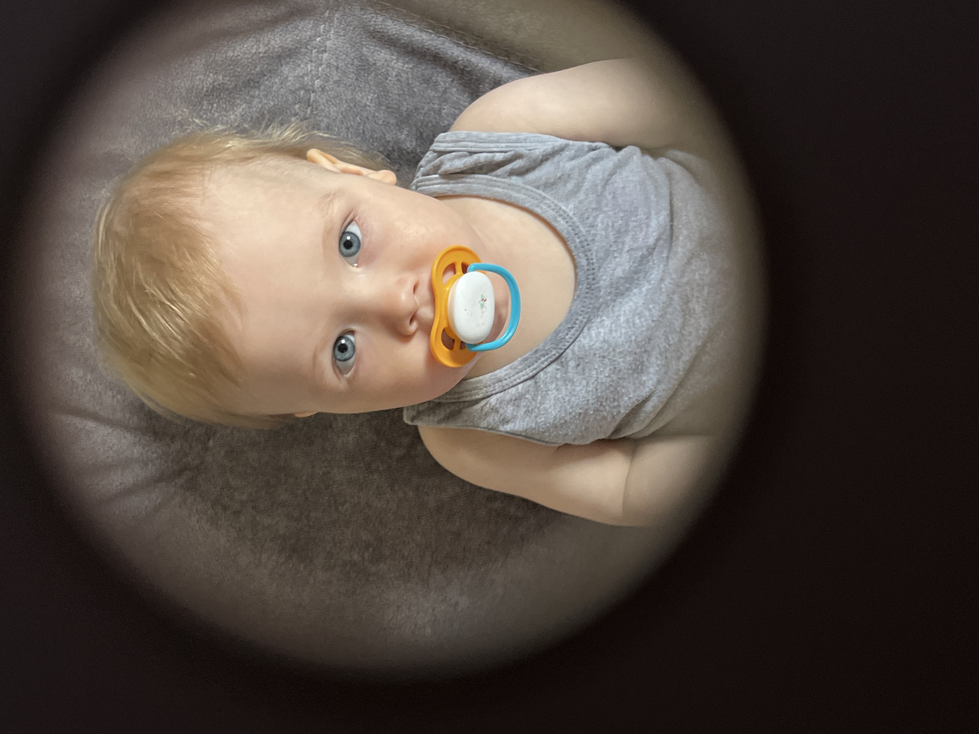 A fair-skinned infant with blond hair and blue eyes lies on a gray, textured surface while wearing a gray sleeveless shirt. The infant has a pacifier with an orange guard and blue handle in its mouth. The image is framed by a dark vignette. There is no information available from the image to determine a location in Chaikovsky, Russia.