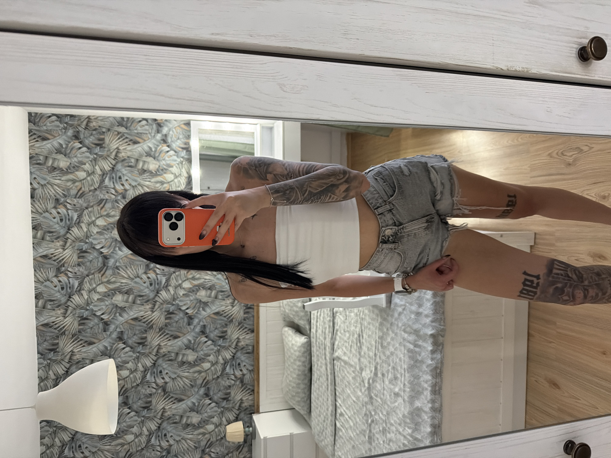 A young woman with black hair is standing in a bedroom and taking a mirror selfie with her orange phone. She is wearing a white tube top and ripped denim shorts. She has tattoos on her arms and legs. The bedroom has a bed with grey patterned bedding, a nightstand with a lamp, and floral wallpaper. The room appears to be located in a residential setting, possibly an apartment or house. There is no indication that this location is in Plovdiv, Bulgaria.