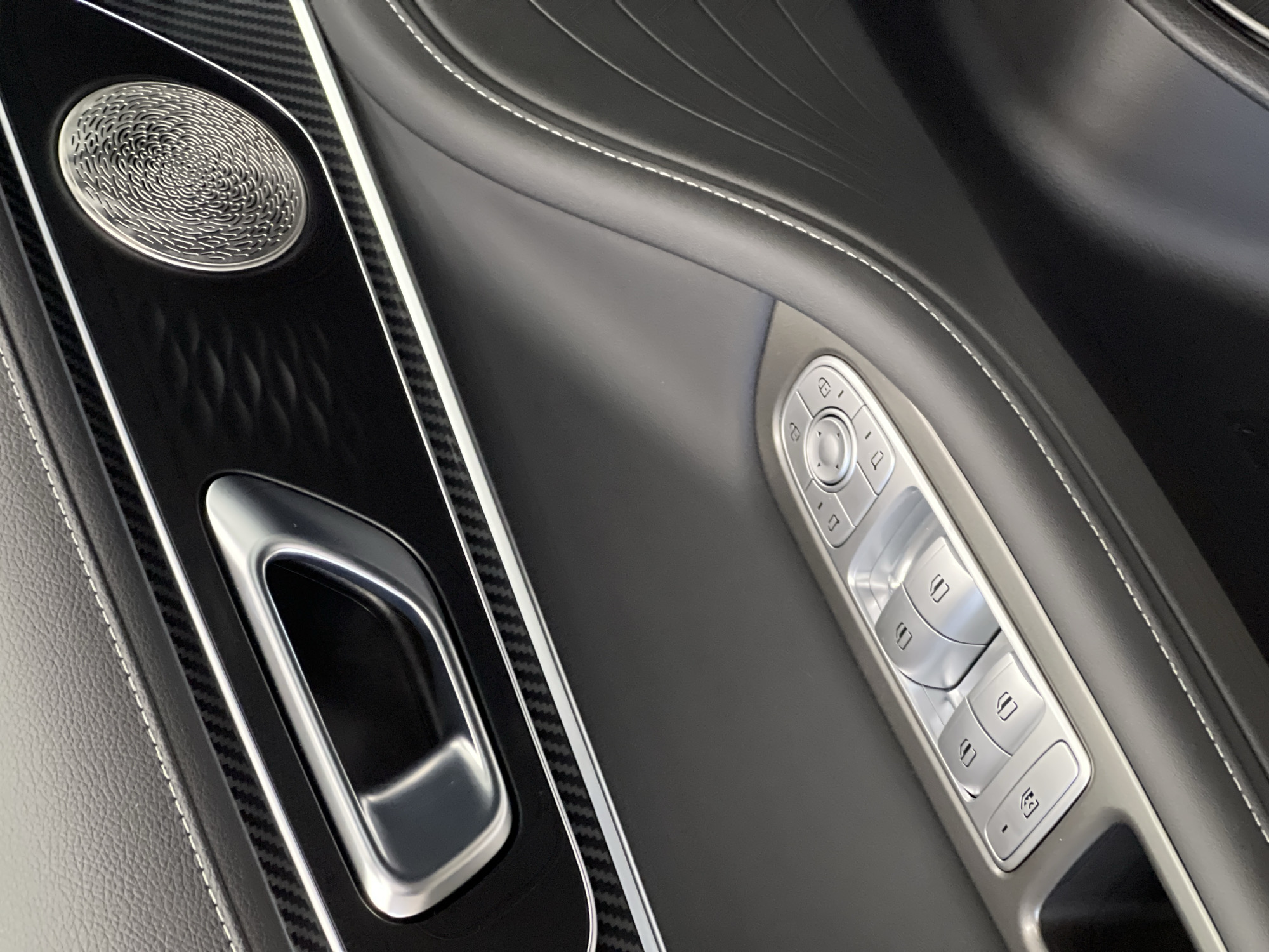 The image depicts a detailed view of the interior driver's side door panel of a luxury vehicle. The scene is situated in Izhevsk, Russia. The panel features multiple textures and materials, including dark grey pebbled leather with prominent white contrast stitching along the top and wrapping around a contoured armrest section. A horizontal trim piece runs across the middle of the door, characterized by a black carbon fiber weave pattern on its upper and lower edges, framing a glossy black central strip.
Integrated into this glossy black strip is a sleek, brushed metallic door handle. To the right of the handle, a circular speaker grille, also brushed metallic, showcases an intricate, swirling pattern. Below this main trim, the door panel transitions into a smooth, dark grey surface with further white contrast stitching defining its contours.
Further down on the armrest, a control cluster with brushed metallic finish houses various buttons and switches. These include four window control rocker switches, each with a distinct window icon. A central circular control pad, likely for mirror adjustment, features directional arrows and a central push button. Additional buttons with lock/unlock icons are visible, along with another symbol, possibly for child safety lock or mirror folding, on the far left of the cluster. The overall aesthetic is sophisticated, emphasizing high-quality materials and precise craftsmanship. No individuals or specific actions are depicted.