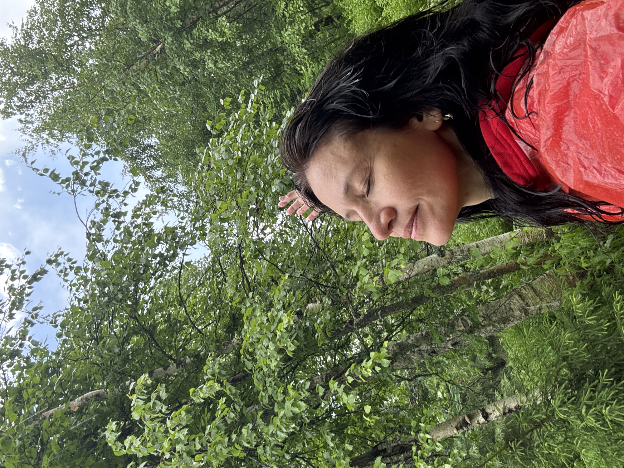 A medium shot captures a woman in a red raincoat with her eyes closed and a slight smile on her face, her head tilted upwards as if enjoying the rain. Her dark, wet hair is spread around her shoulders, and her right hand is raised, palm open, towards the sky. She is surrounded by lush green trees and foliage, with the overcast sky visible through the branches. The setting appears to be a forest or park. The image does not contain specific details to confirm the location as Shumikhinskii, Russia.