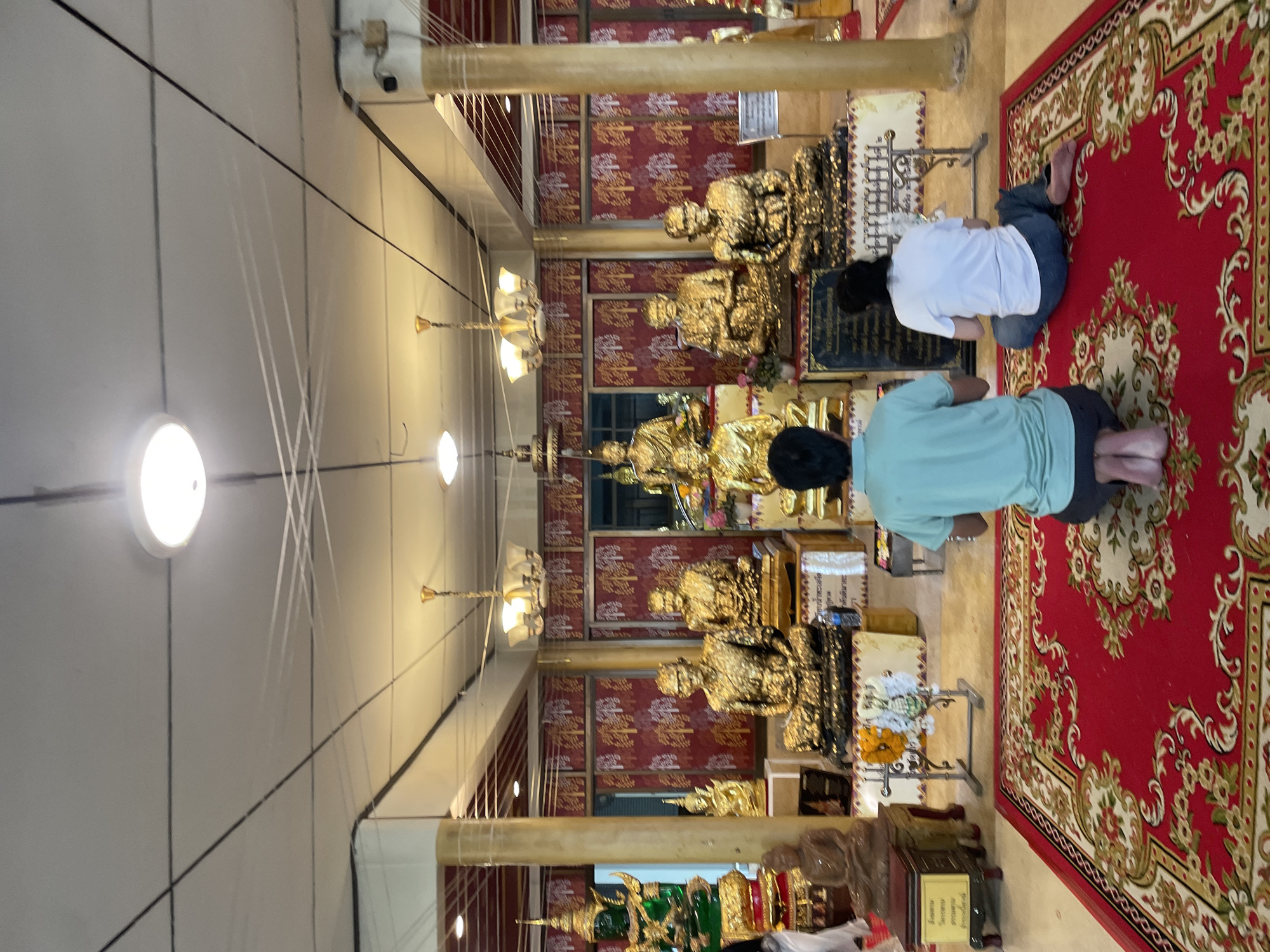 The image shows the interior of a Buddhist temple in Bang Saray, Thailand. Two individuals are kneeling on a red rug in front of a display of golden Buddha statues. The man on the left is wearing a teal shirt and dark shorts, with his knees folded beneath him. The woman on the right is wearing a white t-shirt and blue pants, with her legs extended to the side. The statues are arranged on elevated platforms, with some adorned with intricate gold leaf. The walls are decorated with red fabric featuring a repetitive tree motif. The ceiling is a light color with visible fluorescent lighting and overhead wires. The overall lighting suggests an indoor environment.