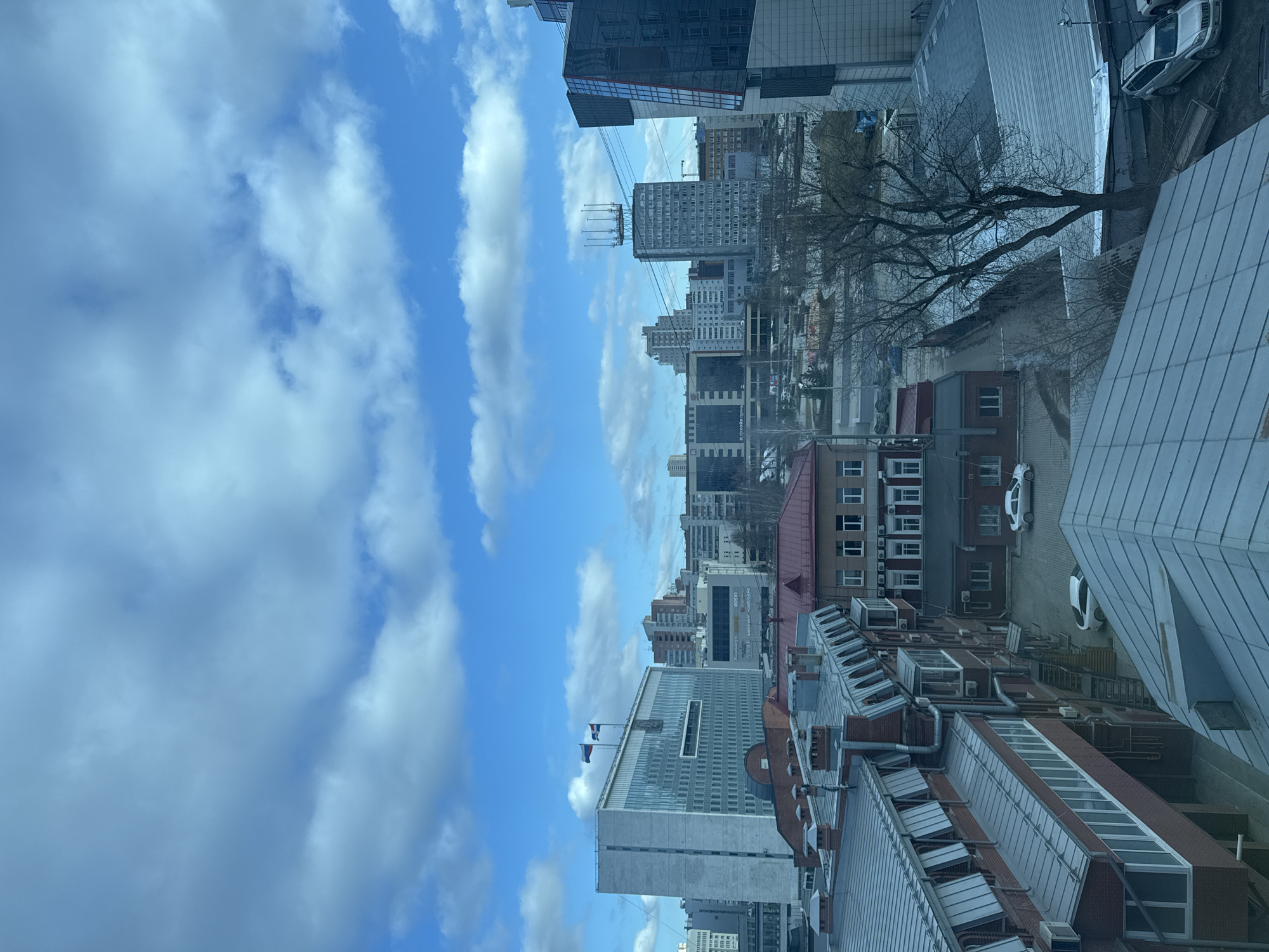 The image depicts a cityscape in Perm, Russia, viewed from an elevated perspective, likely from a high-rise building. The scene is divided between the sky and the urban environment. Above, a blue sky is filled with white cumulus clouds. Below, various buildings dominate the view. There are several high-rise buildings, some with reflective glass facades, amidst a collection of older, brick and concrete structures with red or gray roofs. A large tree with bare branches is visible among the buildings. Several white cars can be seen parked along the rooftops. A large building is visible with two flags flying. The lighting suggests it is daytime.