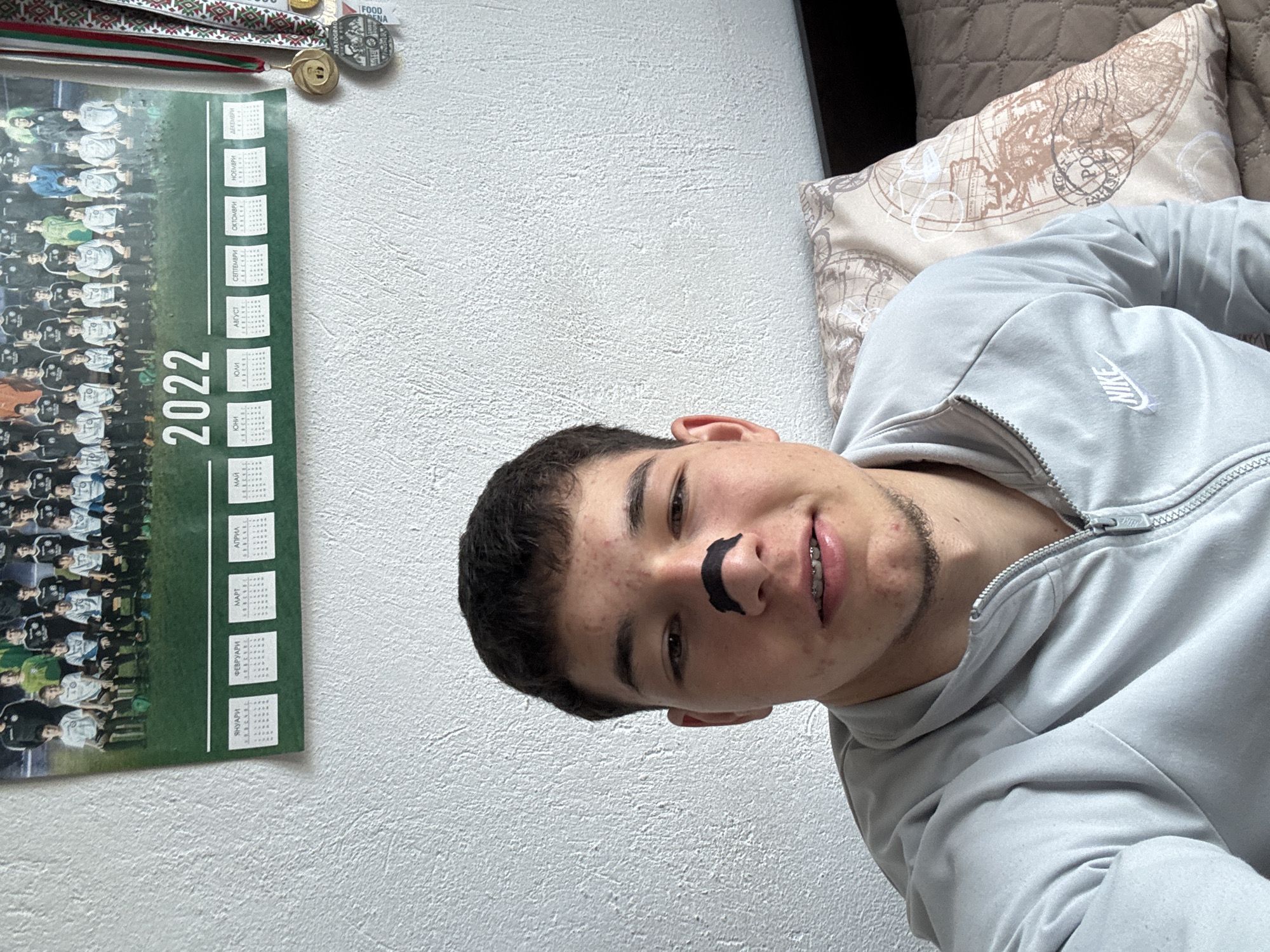 Here is a detailed description of the image:
A young man with dark hair and a fake black mustache is lying on a bed with a patterned pillow. He is wearing a gray Nike hoodie. On the wall behind him is a 2022 calendar in Bulgarian, featuring a sports team photograph. Above the calendar are several medals and a ribbon in the colors of the Bulgarian flag. The wall is textured. The presence of a calendar in Bulgarian suggests the location is likely in Bulgaria. The presence of "Z" and "FOOD" on the medal suggests that the location is the city of Zlatograd.
