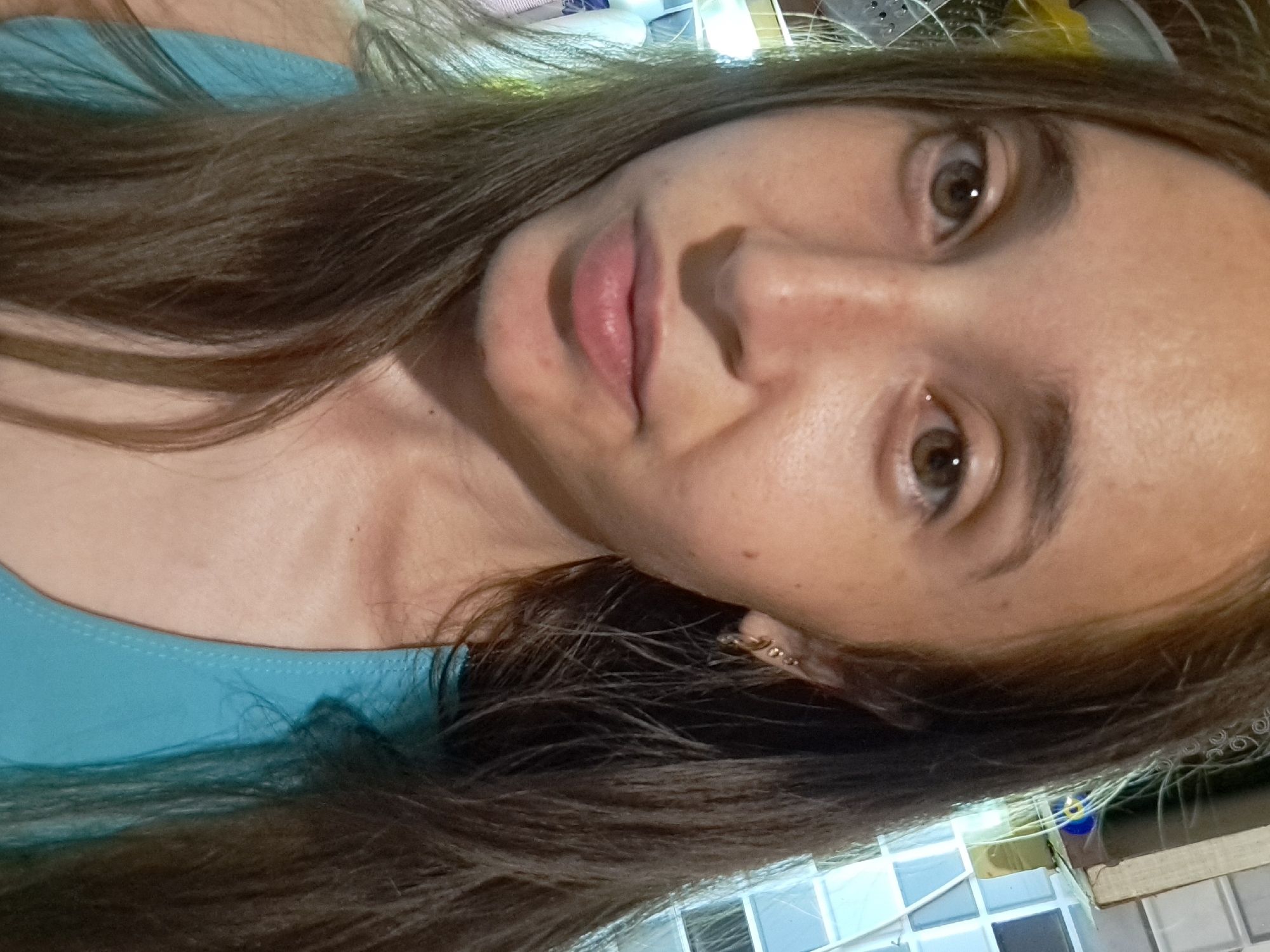 A close-up, forward-facing selfie of a young woman with long, dark brown hair, fair skin, and brown eyes. She is wearing a light blue tank top. Her left ear is visible and adorned with a small gold earring. The background is blurred and appears to be indoors, with a tiled wall visible behind her head. The image was taken in Vypasne, Ukraine.