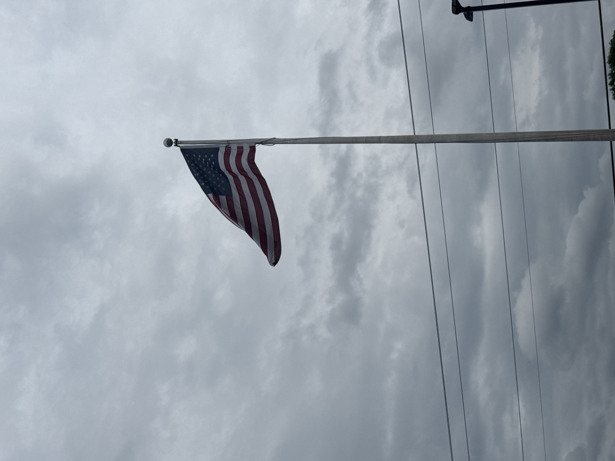 The image shows an American flag attached to a flagpole, flying against a cloudy sky. Power lines run vertically across the right side of the frame. Some greenery is visible in the upper right corner. The location is the city of Stone Mountain, United States of America.