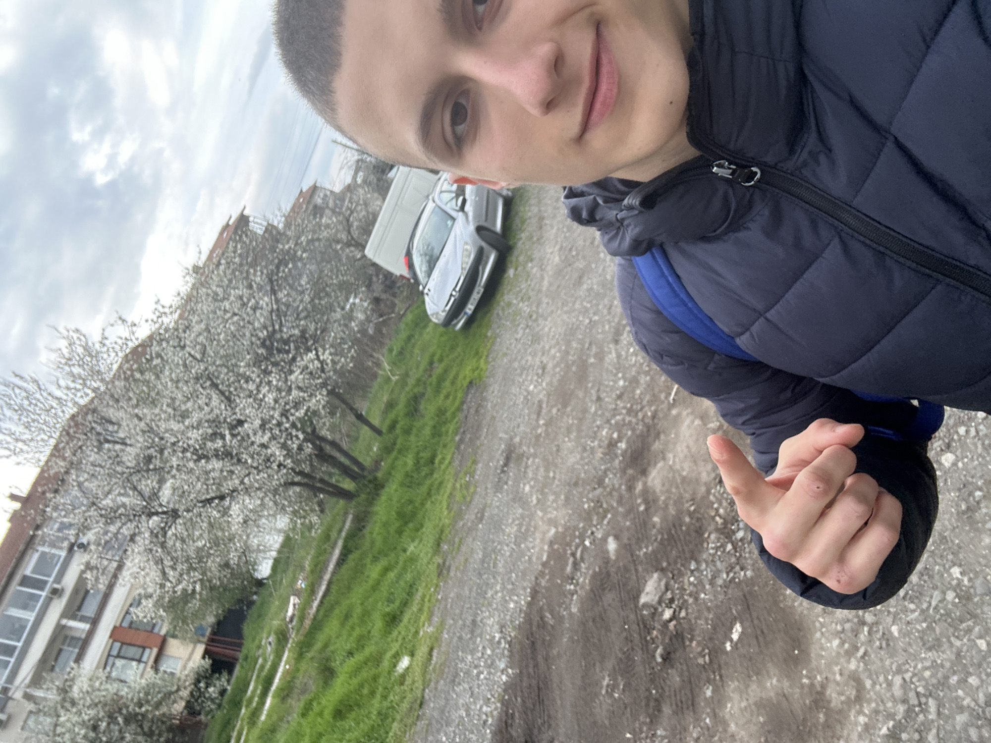 A young man is taking a selfie. He has short, dark hair, light skin, and is wearing a navy blue quilted jacket and a blue backpack. He is holding up his left hand, appearing to give the "hang loose" sign. Behind him, there is a building with white walls and multiple windows. A tree with white blossoms is in full bloom to the left of the building. In the background, two vehicles are visible, including a silver car and a white van. The ground is a mix of gravel and patches of green grass. The sky is overcast. The location appears to be a residential area or a parking lot. It is uncertain from the visual data whether this photo was taken in Burgas, Bulgaria.