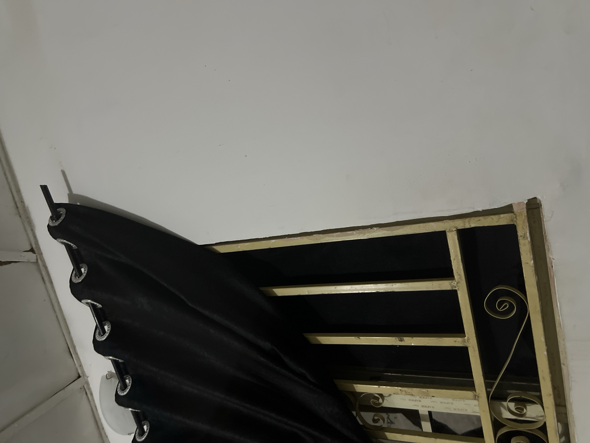 A black curtain with grommets is partially covering a window with a gold metal grate. The curtain is drawn to the left, revealing bars of the grate and a dark interior. The wall on the right is painted white and shows some minor imperfections. The scene is indoors, as suggested by the ceiling structure visible at the top and the presence of a window with a protective grate. The image does not provide any information that would indicate its location within Howayi, Nigeria, or any specific context beyond the visual elements. There are no people or actions depicted.