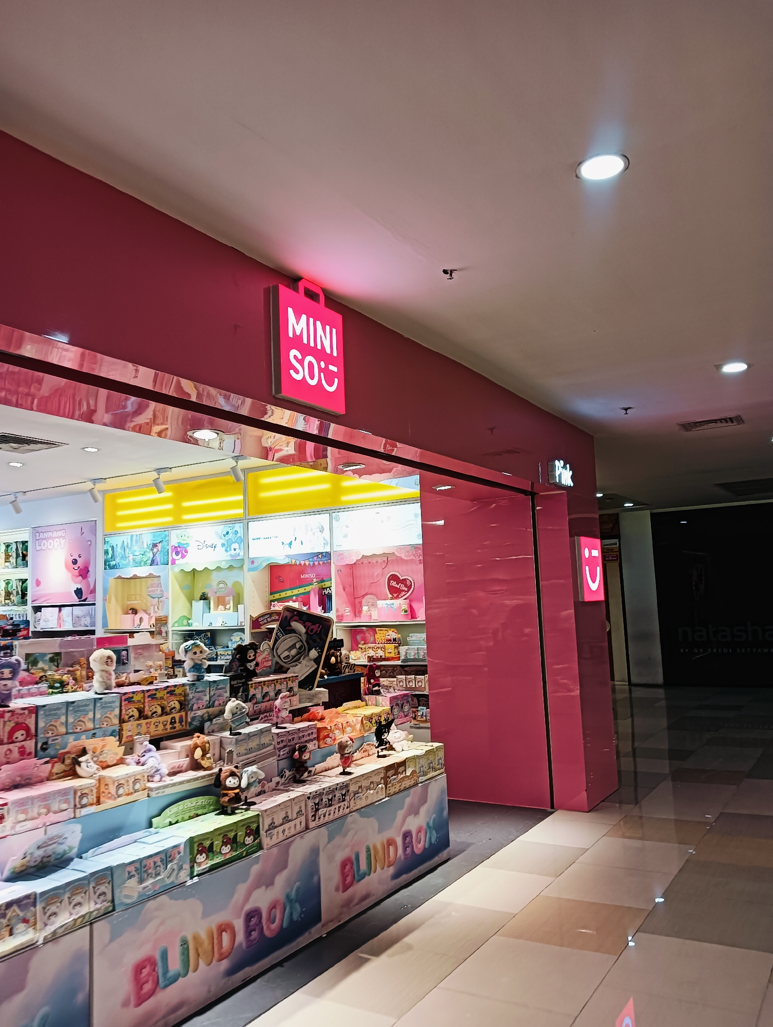 The image depicts the interior of a shopping mall in Batam, Indonesia, specifically focusing on a brightly lit Miniso store. The store's facade is predominantly pink, with a prominent neon sign displaying "MINI SO" in white and a red shopping bag logo. Inside, shelves are filled with various merchandise, including plush toys, blind boxes, and decorative items, presented in a colorful and organized manner. The flooring is tiled, reflecting the overhead lighting. In the background, another store with the name "natasha" is visible, along with other commercial spaces and mall architecture. The overall impression is one of a modern retail environment.
