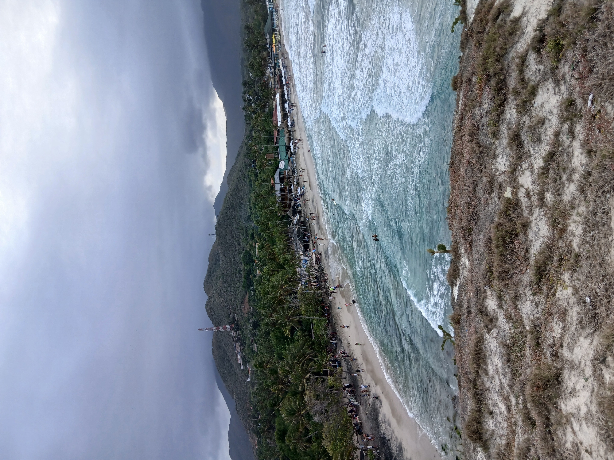 The image shows a beach scene, possibly taken from an elevated position. The sky is overcast with heavy clouds. In the distance, there are mountains partially obscured by the clouds. Closer to the beach, lush green vegetation and palm trees line the coast, suggesting a tropical or subtropical climate. A strip of buildings is visible along the beach. People are present on the beach, walking along the shore or swimming in the water. The ocean has strong waves, indicating potentially rough sea conditions. On the right side of the frame, there is an area of dry, sandy vegetation. A tall communication tower can be seen near the buildings. The location is Unknown, Unknown.