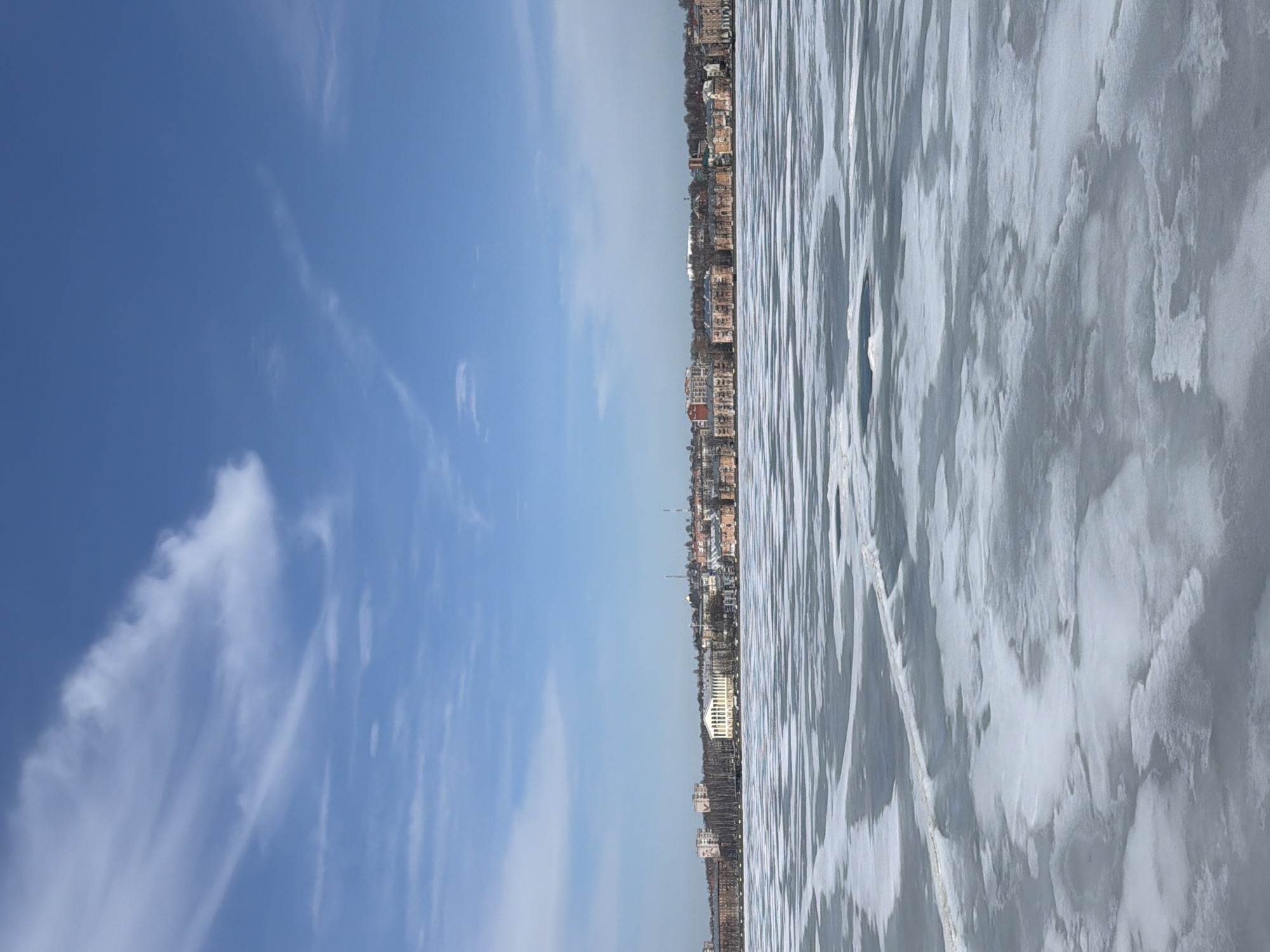 The image depicts a waterfront scene in Votkinsk, Russia. On the left side, a clear blue sky with wispy clouds is visible. The right side shows a frozen body of water, likely the Votkinsk Reservoir, with a textured, icy surface. In the middle ground, a city skyline with various buildings and structures stretches horizontally. The buildings appear to be a mix of residential and commercial properties, ranging in height and design. The frozen water suggests a cold, possibly winter, season. No people are visible in the image.