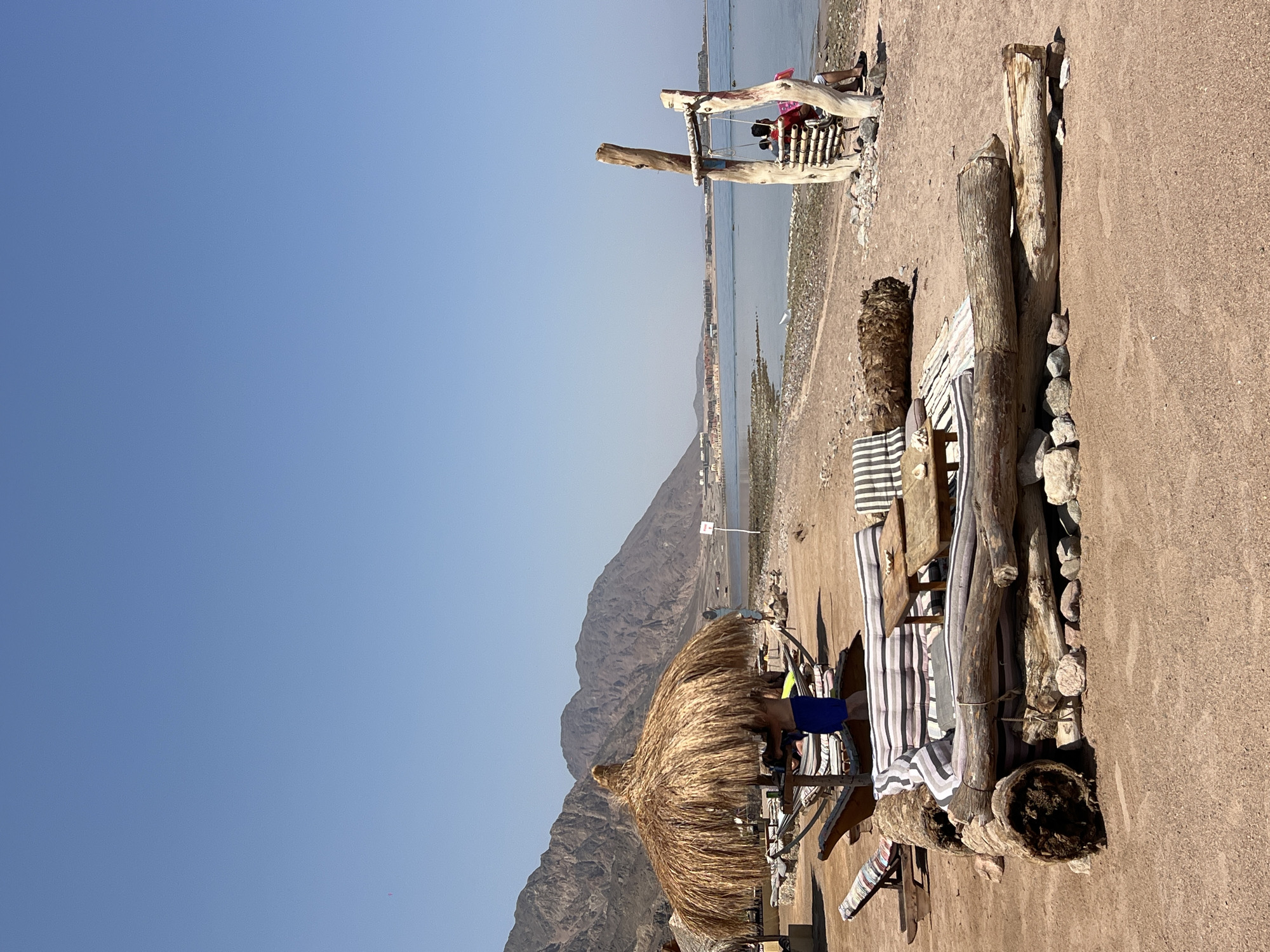 This image depicts a beach scene in an unknown location in Egypt. The foreground is dominated by a rustic seating area constructed from logs and adorned with striped cushions and a small wooden table. To the left, a person in blue shorts stands near a thatched umbrella, with lounge chairs and hammocks visible. In the middle ground, the sandy beach gives way to a calm body of water, with mountains forming a backdrop. Further down the shoreline, a wooden swing with two occupants is situated near the water's edge. Buildings are visible in the distance along the coast. The sky is clear and blue.