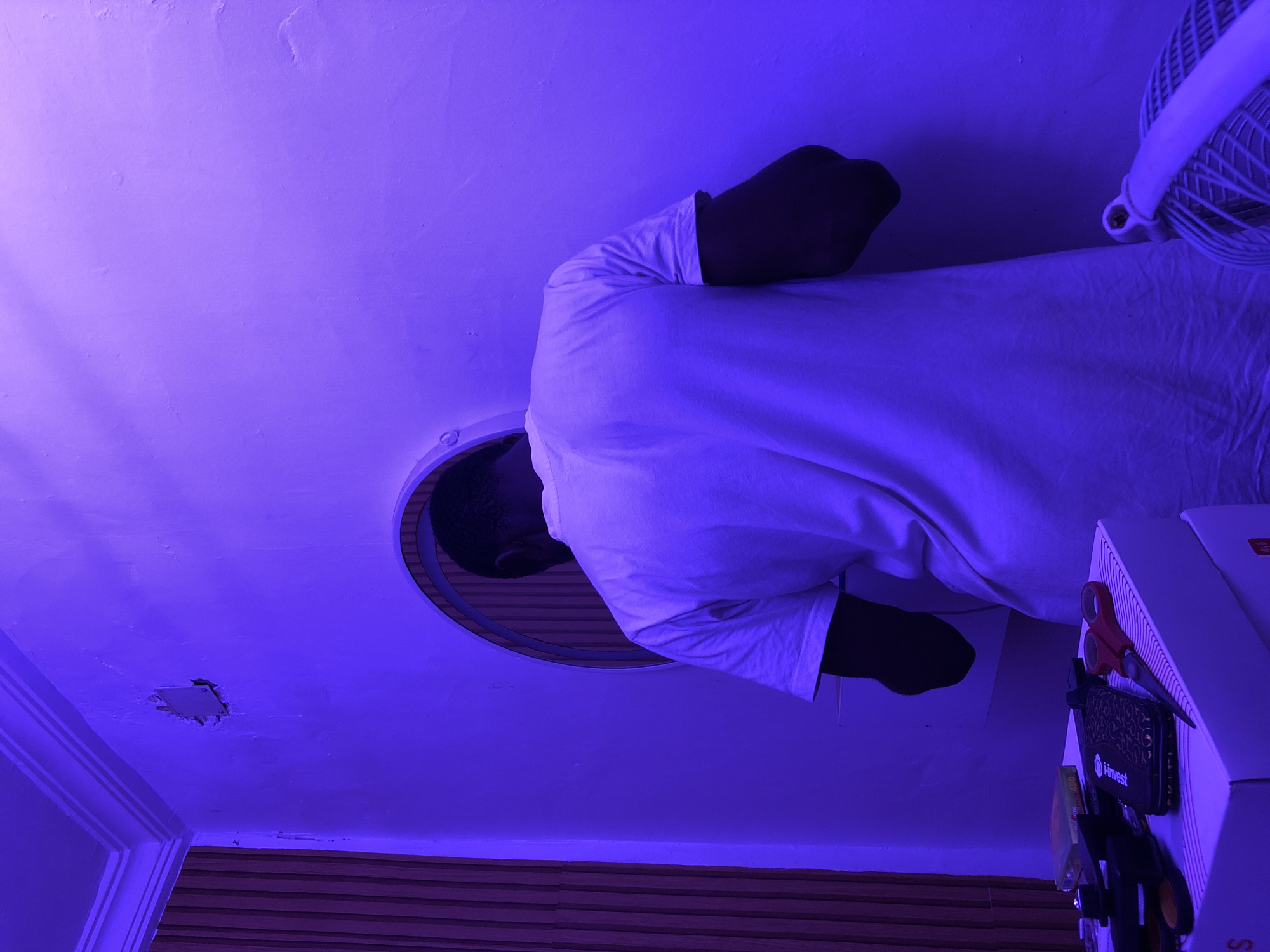 A person lies in a bed under a purple light. The person wears a white shirt. There is a circular mirror and a white fan on the ceiling. Other objects include what appears to be scissors, a black wallet, and a white box on a nearby surface. The exact location is unknown.