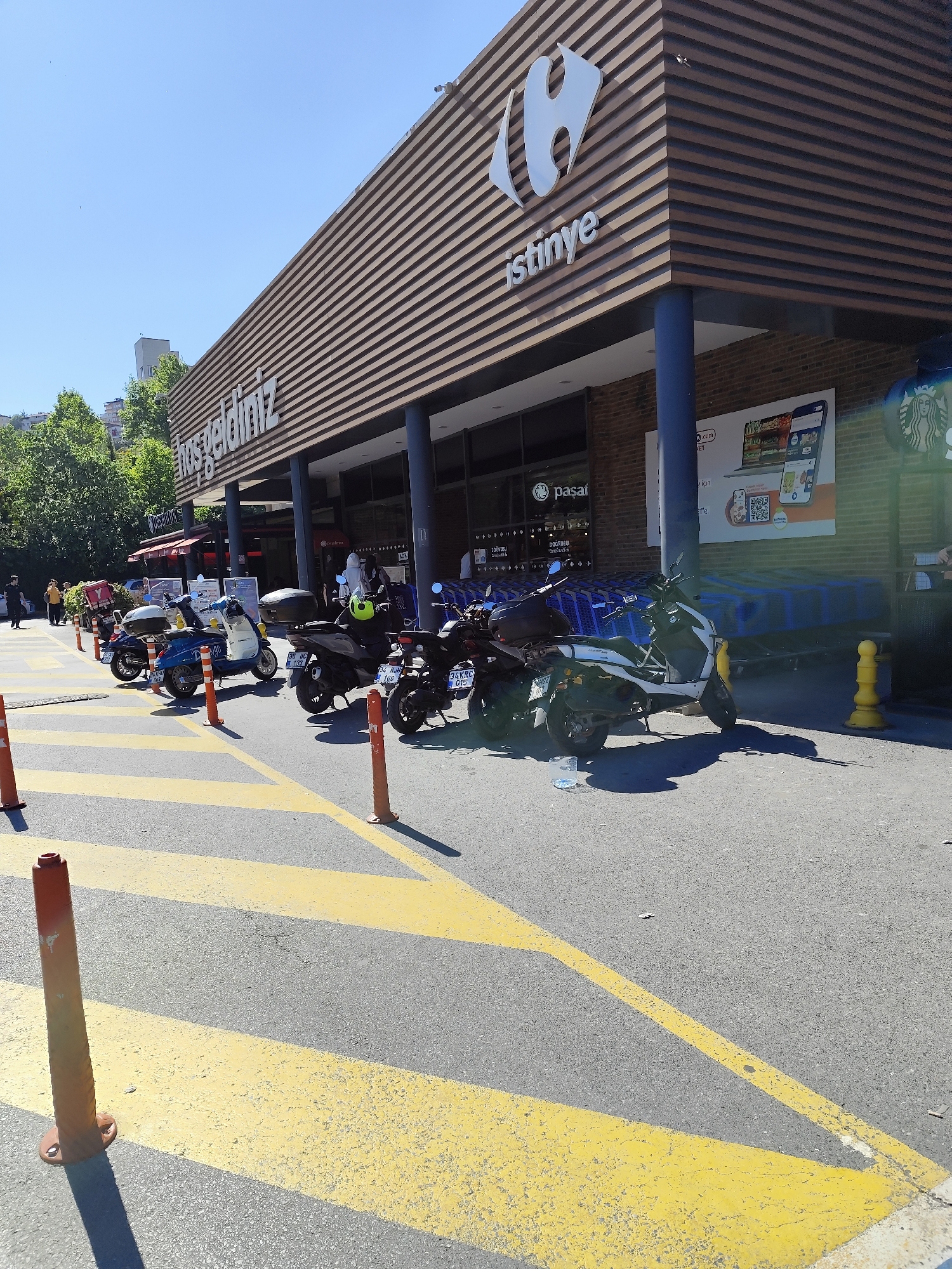 The image captures a row of parked motorbikes in front of a Carrefour SA supermarket located in the Istinye neighborhood of Sarıyer, Turkey. The building is distinguished by its brown horizontal striped facade, with the "istinye" brand and Carrefour's logo displayed prominently above the entrance. Underneath the awning, a Starbucks logo can be seen. In the foreground, yellow striped pedestrian crosswalks and orange traffic poles delineate the parking area. To the left, a group of trees is visible. A number of people are present in the image. The sky is clear and blue, indicating fair weather conditions.