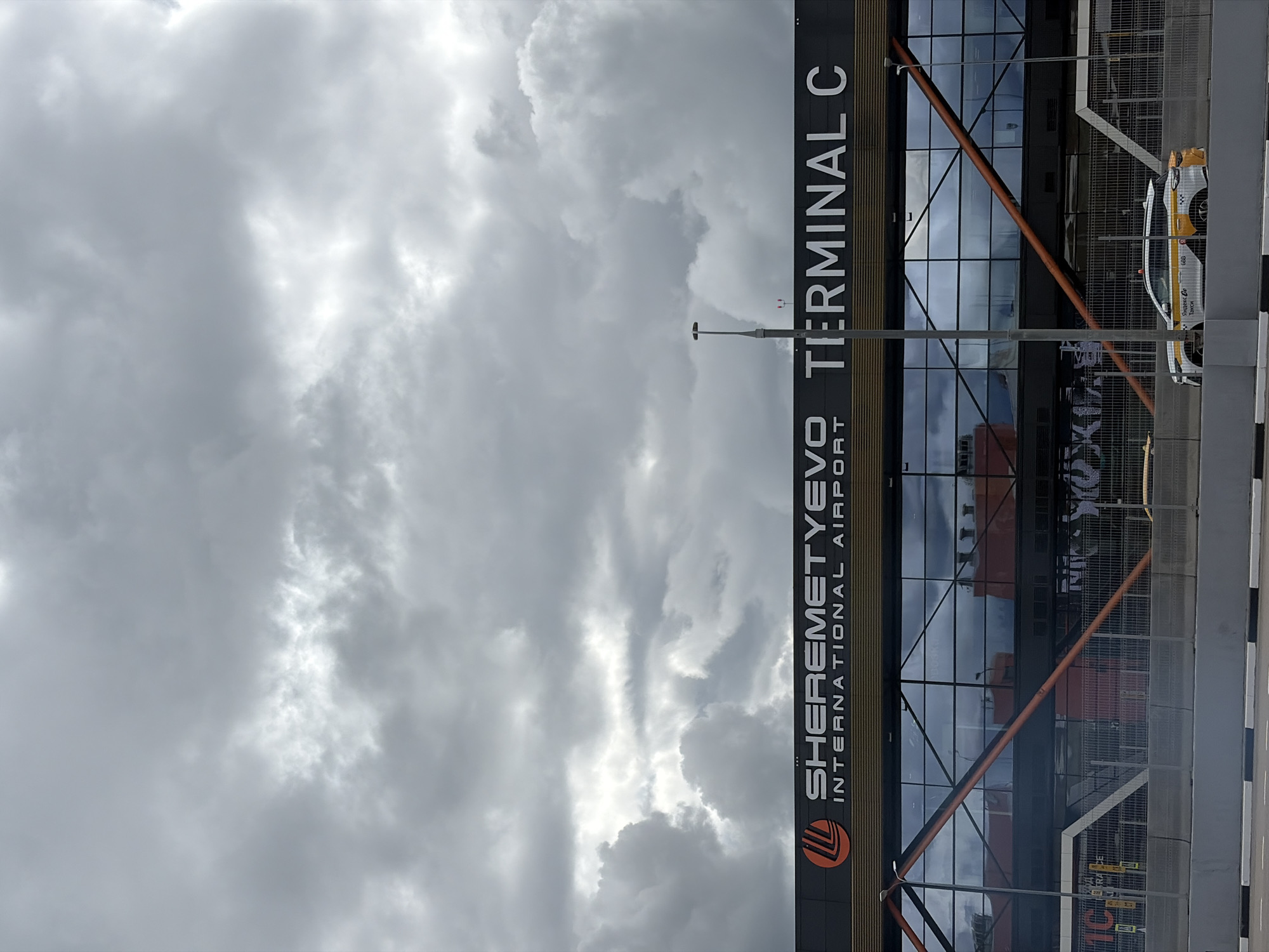 The image shows the exterior of Terminal C at Sheremetyevo International Airport in Khimki, Russia. The terminal building is visible, featuring a modern design with large windows. The signage "SHEREMETYEVO INTERNATIONAL AIRPORT TERMINAL C" is prominently displayed on the building's facade. In the foreground, a security fence and roadway are visible. A yellow and white taxi is parked along the roadway. The sky is overcast with gray clouds.