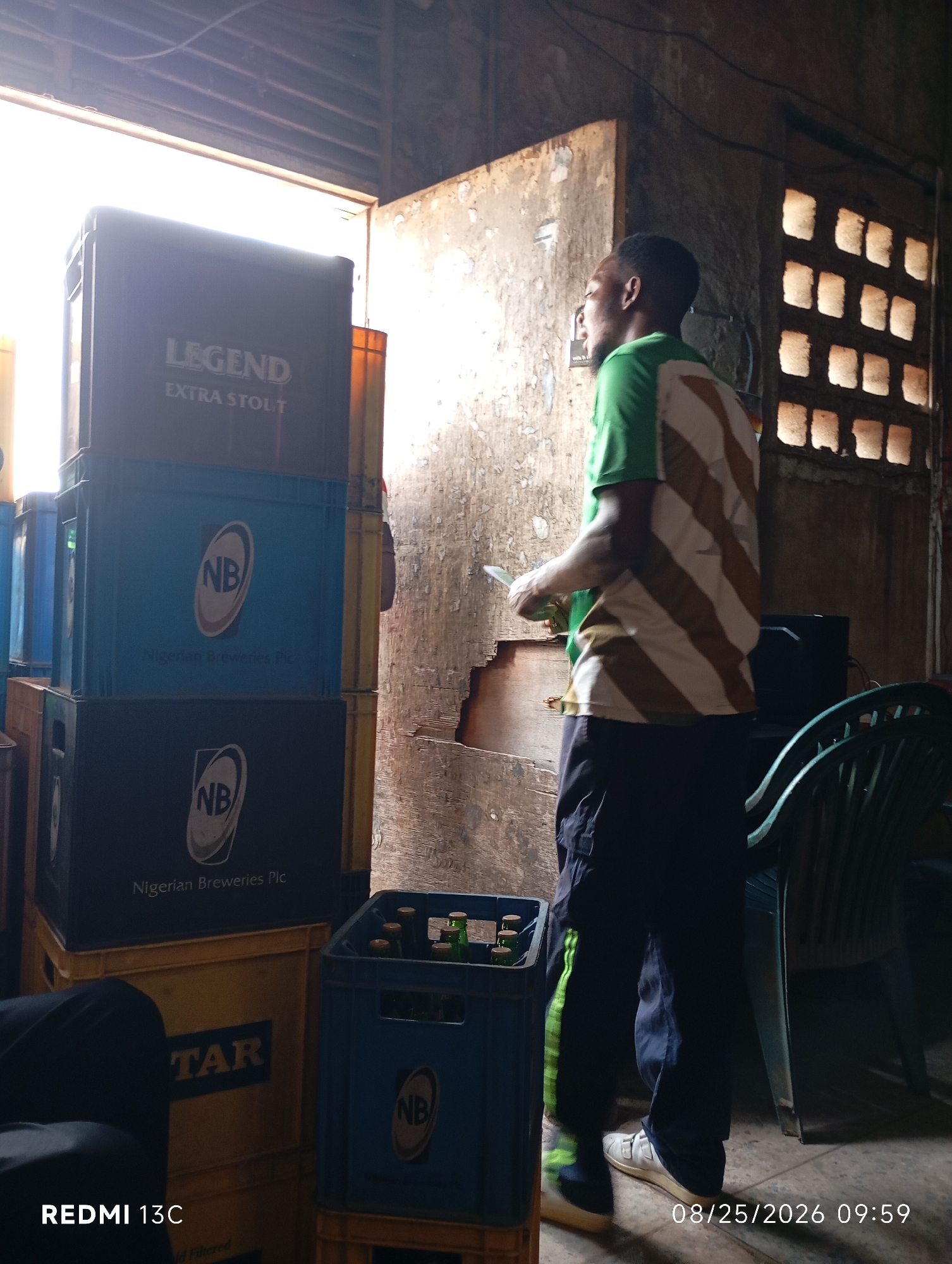 A young Black male, wearing a green and brown striped t-shirt and dark pants with green stripes, stands in an indoor space that appears to be a storage area or informal shop. He is facing away from the camera, looking towards a doorway or open space illuminated by bright sunlight. In his right hand, he holds a silver object, possibly a knife or a tool.
The environment is rustic and appears to be in a state of disarray. The walls are made of rough, weathered wood or plaster. Natural light streams in from an open doorway and through a grid of square windows or vents in the background, casting strong highlights and shadows. Stacks of plastic crates, predominantly dark blue, black, and yellow, are visible throughout the scene, some filled with bottles. The crate at the front right contains several green bottles with brown caps. A green plastic chair is positioned to the right of the male.
The text "LEGEND EXTRA STOUT" is visible on a dark crate at the top left. Below it, "NB" and "Nigerian Breweries Plc" are imprinted on a blue crate. A yellow crate with the text "STAR" is partially visible. The bottom right corner of the image displays a timestamp: "08/25/2026 09:59" and "REDMI 13C". The context suggests this scene is located in Shendam, Nigeria, likely a local business or distribution point.
