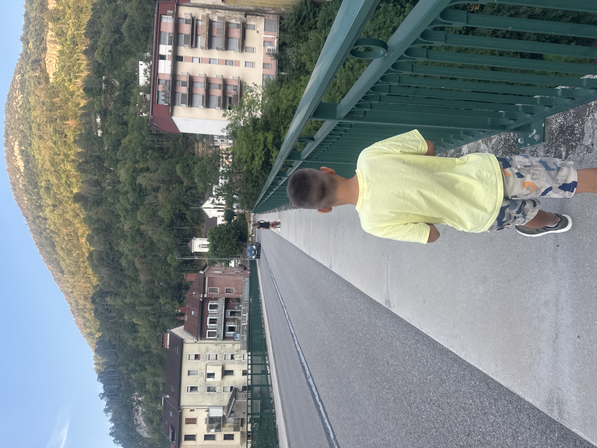 An over-the-shoulder shot depicts a young boy walking on a sidewalk beside a road on a bridge in Saint-Claude, France. The boy wears a yellow t-shirt and camouflage shorts. In the background, buildings line the street, and a tree-covered mountain rises behind them. A dark-colored car is visible on the road. Another person on a bicycle is also present further down the bridge. The sky is clear and blue, suggesting daytime.