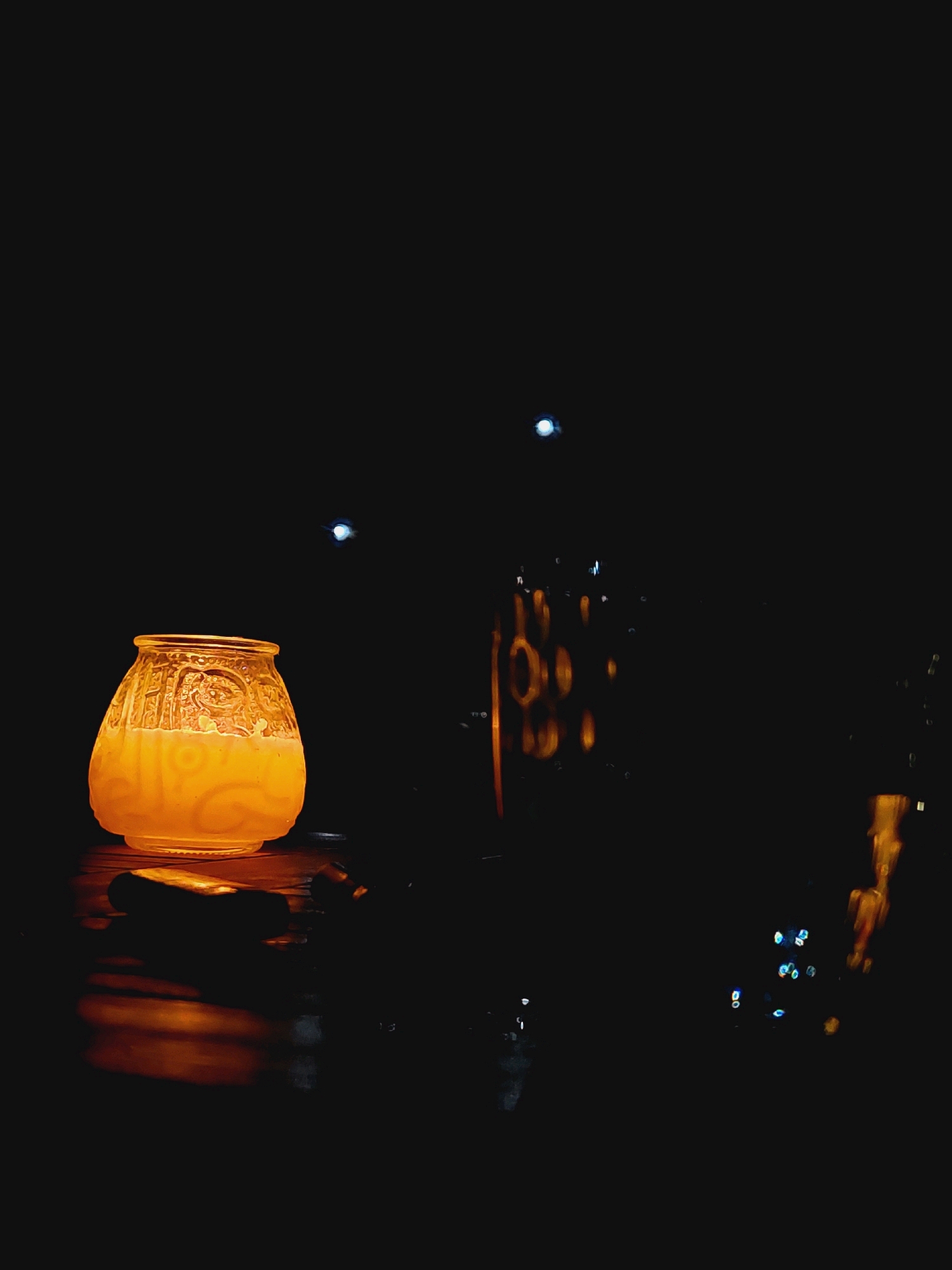 An illuminated glass jar sits on a dark, reflective surface. The jar emits a warm, orange glow and appears to contain a creamy substance. The surface below the jar is wet, reflecting the light from the jar. In the background, out of focus, are distant lights, some appearing as blurred orbs of white and blue. The overall scene is dark, suggesting it is nighttime. The location is identified as Sozopol, Bulgaria.