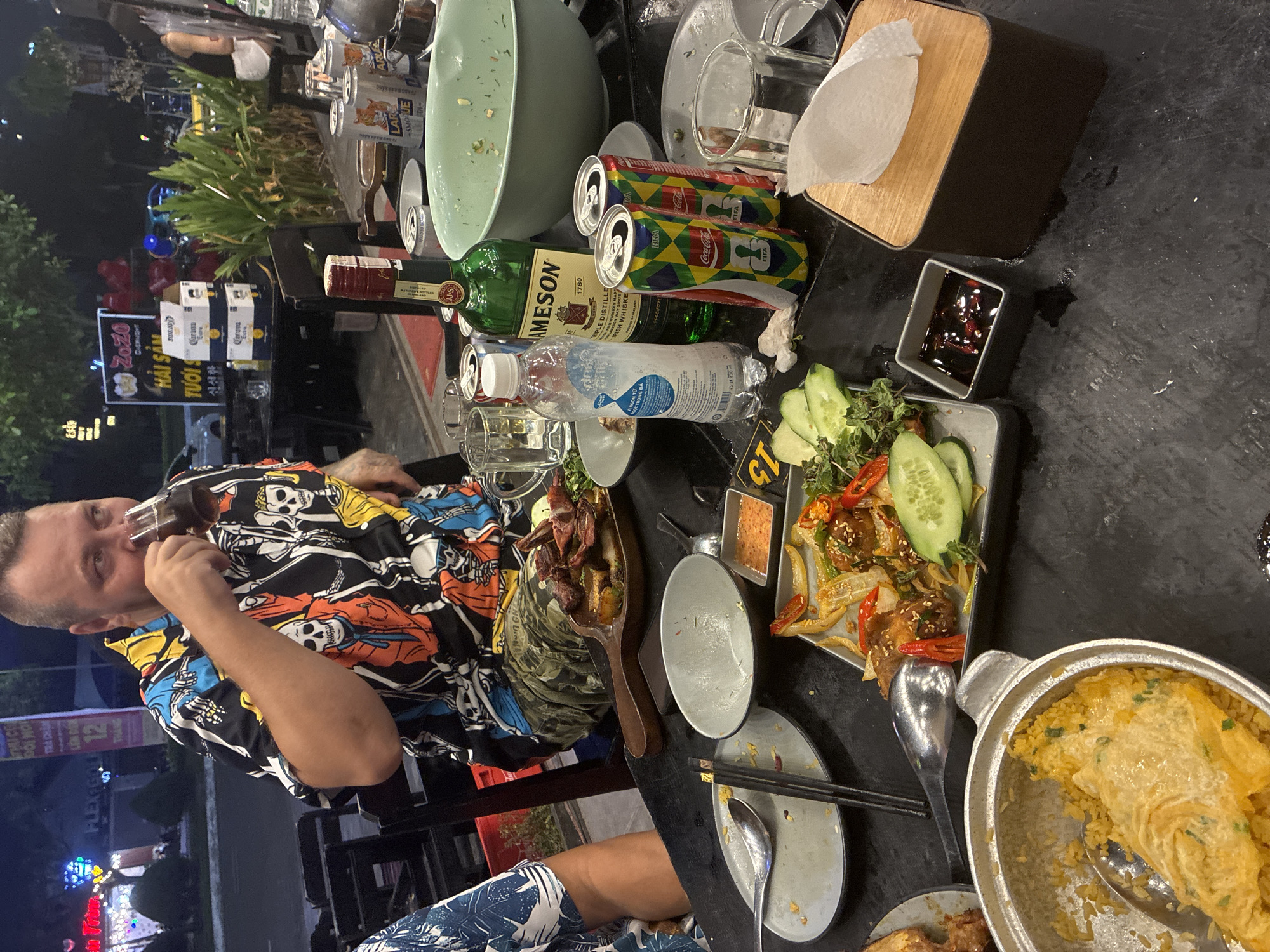 The image depicts an outdoor dining setting in Đà Nẵng, Vietnam. A man wearing a skull-patterned shirt is drinking from a glass. The table is cluttered with various food dishes, including fried rice, meat skewers, and a vegetable plate with cucumbers. There are also empty bowls and plates, as well as Coca-Cola cans, a Jameson bottle, and a bottle of water. A wooden tissue box and a small container of dipping sauce are also present. In the background, there is a sign that reads "Zozo HẢI SẢN TƯƠI" in Vietnamese, indicating that the establishment specializes in fresh seafood. The background also includes street lights and the outline of buildings.