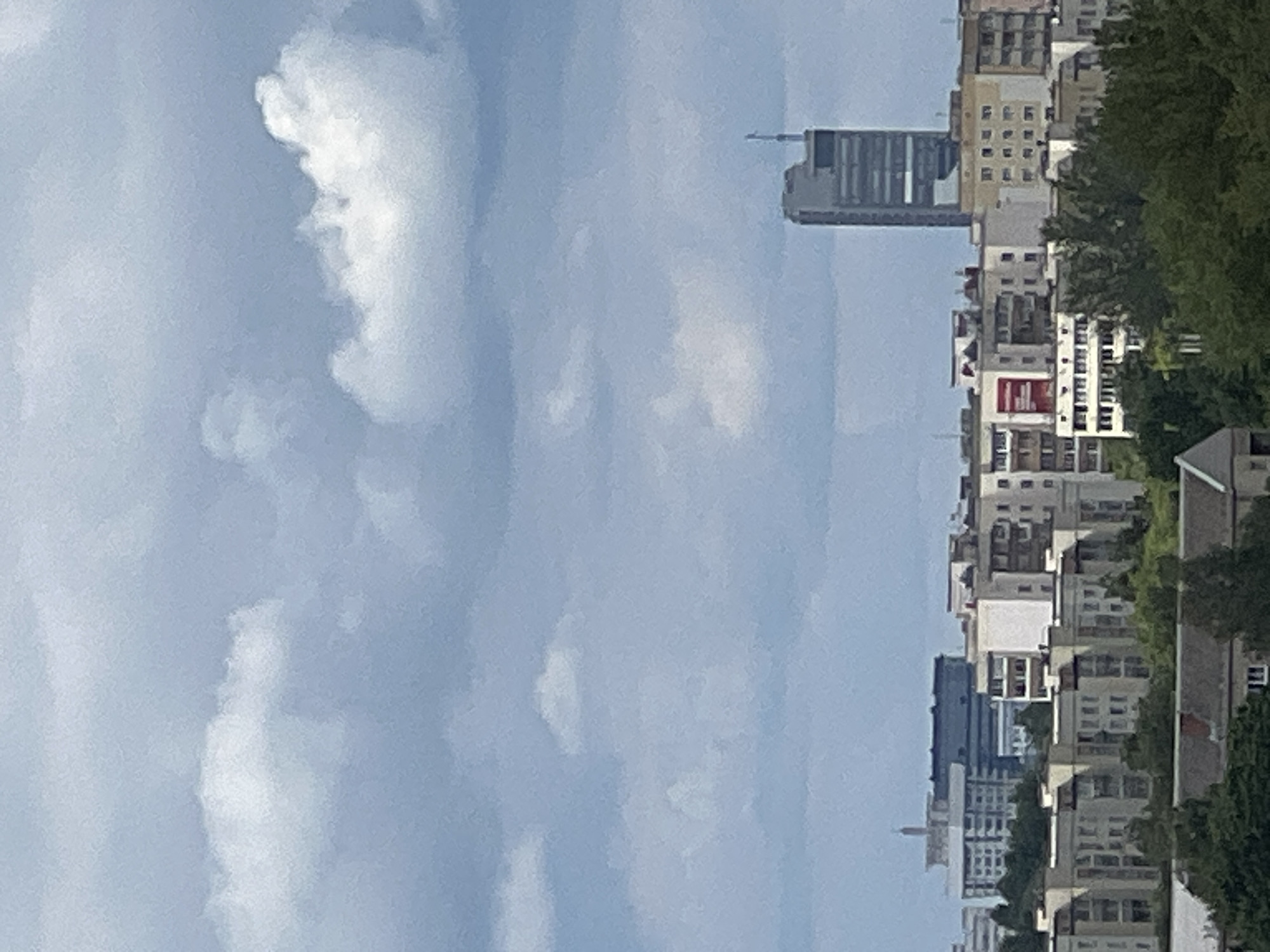 The image shows a cityscape with buildings and clouds. The perspective is vertical, making it appear as if the buildings are extending upwards towards the sky. The sky is a pale blue with scattered white clouds. On the right side of the image, a row of buildings of varying heights is visible, some with windows and balconies. A prominent tall, rectangular building with a darker facade stands out among them. Trees with lush green foliage are interspersed between the buildings and along the side of the scene. The overall impression is that of a bright, possibly sunny day, given the clarity of the sky. The setting appears to be urban, with the density of buildings suggesting a city. No people are visible in the frame. There is some text visible on a red rectangular sign on one of the buildings, but it is not legible. The crops provided offer closer views of the clouds and the buildings, highlighting the architectural details and the textures of the sky.