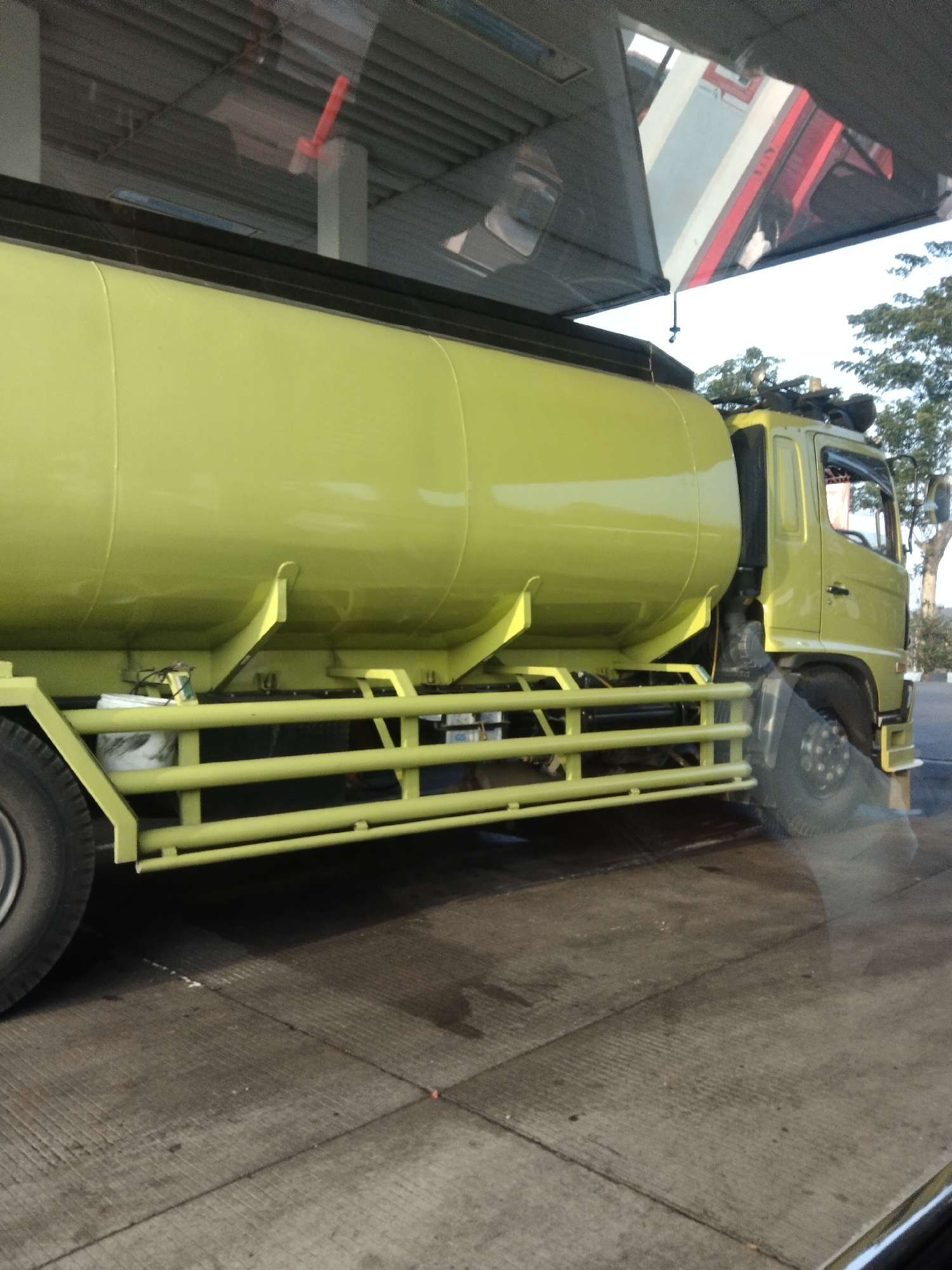 The image shows a side view of a light yellow tank truck parked on a concrete surface. The truck has a long, cylindrical tank. The cab of the truck is visible on the right side, and the front wheel is on the left. There are reflective surfaces on the top of the image, suggesting the photo was taken through a window. In the background, there are trees and a structure with a corrugated roof. The ground appears to be a paved area, possibly a road or a parking lot, with some faint lines suggesting a grid pattern. There is a white bucket with a black lid visible under the tank. The overall scene is of a parked vehicle in an outdoor urban or semi-urban environment.