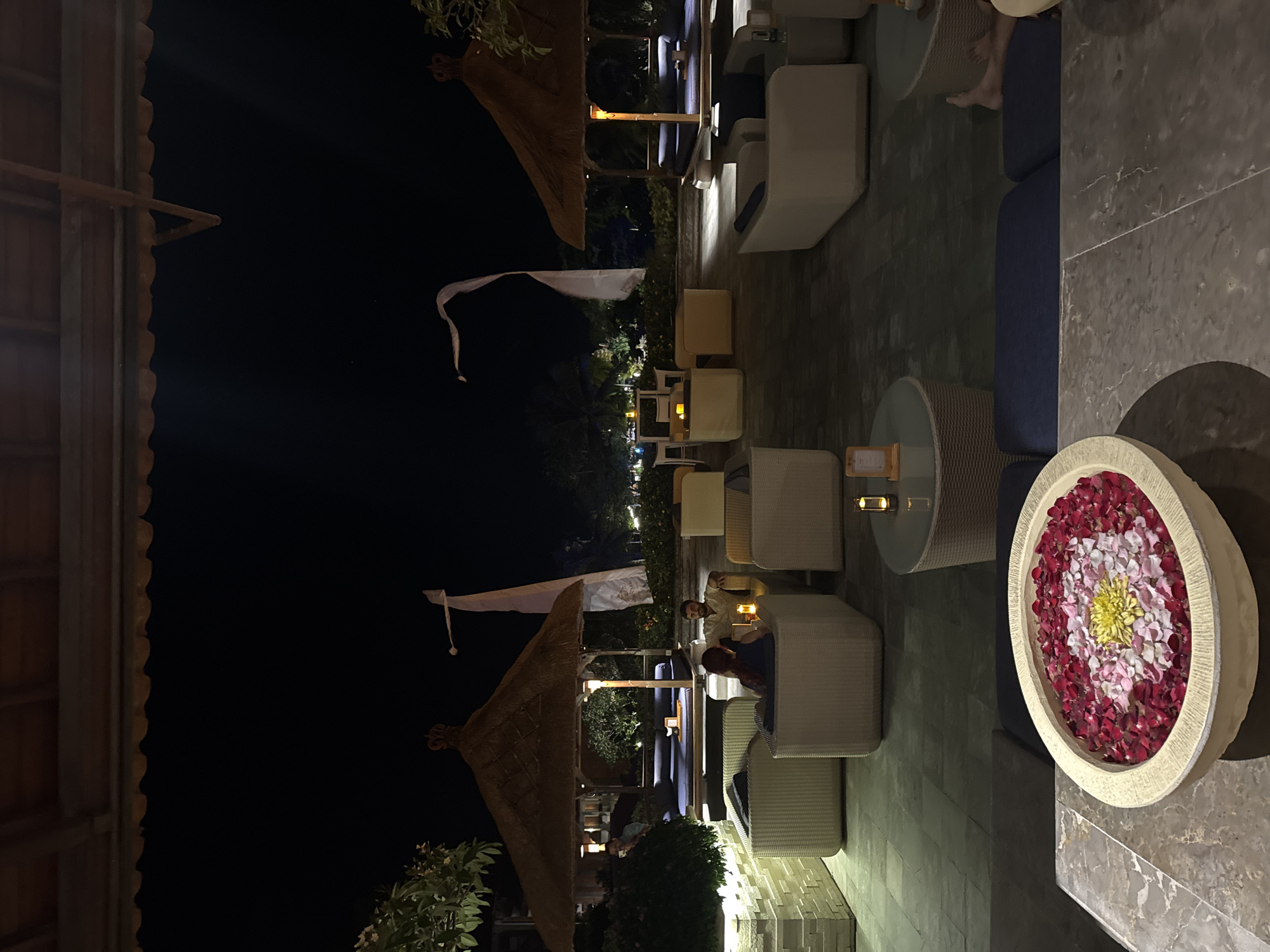 An outdoor seating area at night in Kuta Selatan, Indonesia features a tiled floor, thatched roof gazebos with lounge beds, and white wicker furniture. The scene includes a white bowl filled with red and pink rose petals and a yellow flower in the foreground. Several small tables with candles are scattered throughout the area, and white flags are visible in the background. Two people are seated on a lounge bed within a gazebo, and another person is seated in a wicker chair. The lighting is dim, with yellow-toned lamps illuminating the seating areas.
